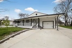 White exterior with dark trim accents, covered front porch with white railings, attached carport, concrete driveway, and an attached garage with a black-framed door
