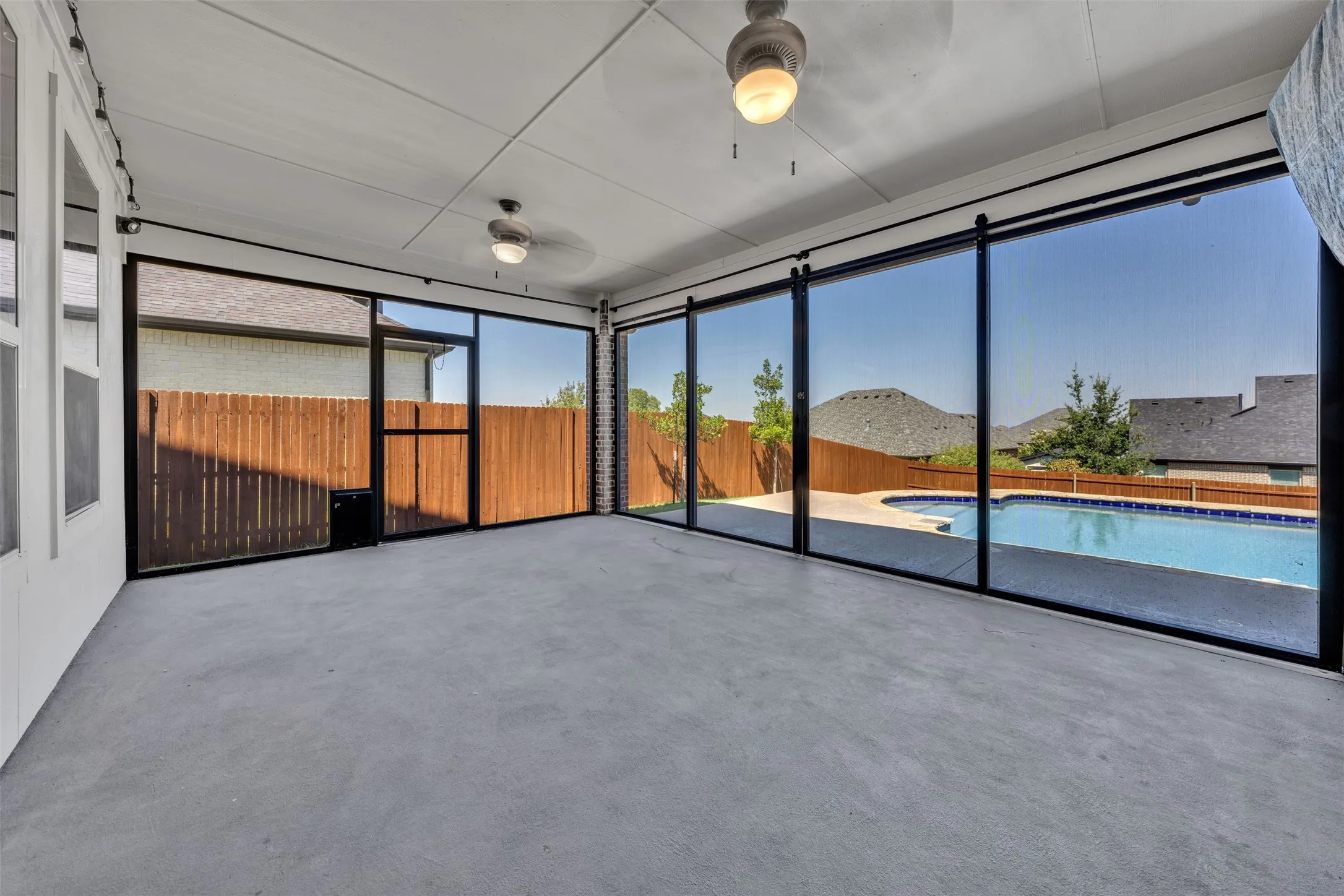 Screened patio with concrete flooring, multiple ceiling fans, and expansive windows overlooking a swimming pool and wood privacy fence
