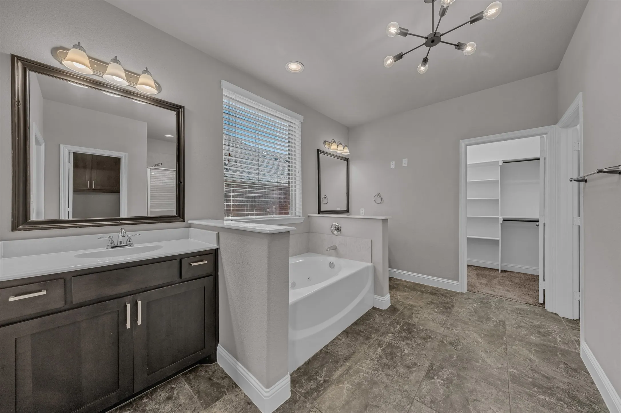Dual vanities with white countertops, a built-in jetted tub, a window with blinds, a modern ceiling light fixture, and a walk-in closet with built-in shelving