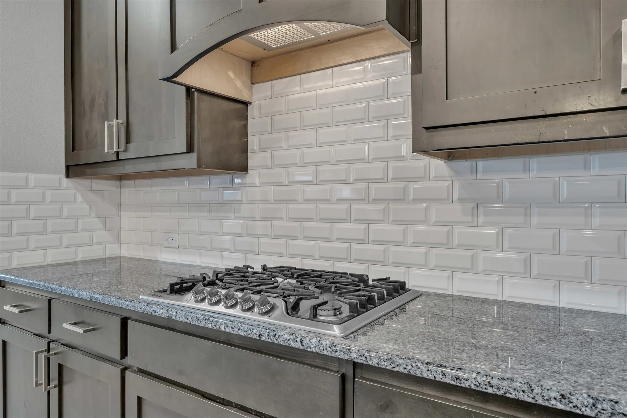 Built-in gas cooktop, granite countertops, subway tile backsplash, custom wood cabinetry, and a range hood