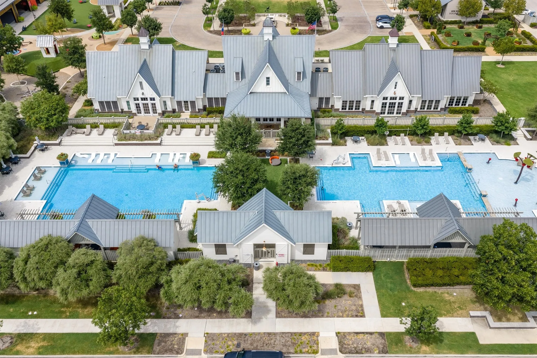 Welcome to Pecan Square Community clubhouse with gabled metal roofs, expansive swimming pool, separate children's splash pad, surrounding lounge areas, and mature landscaping