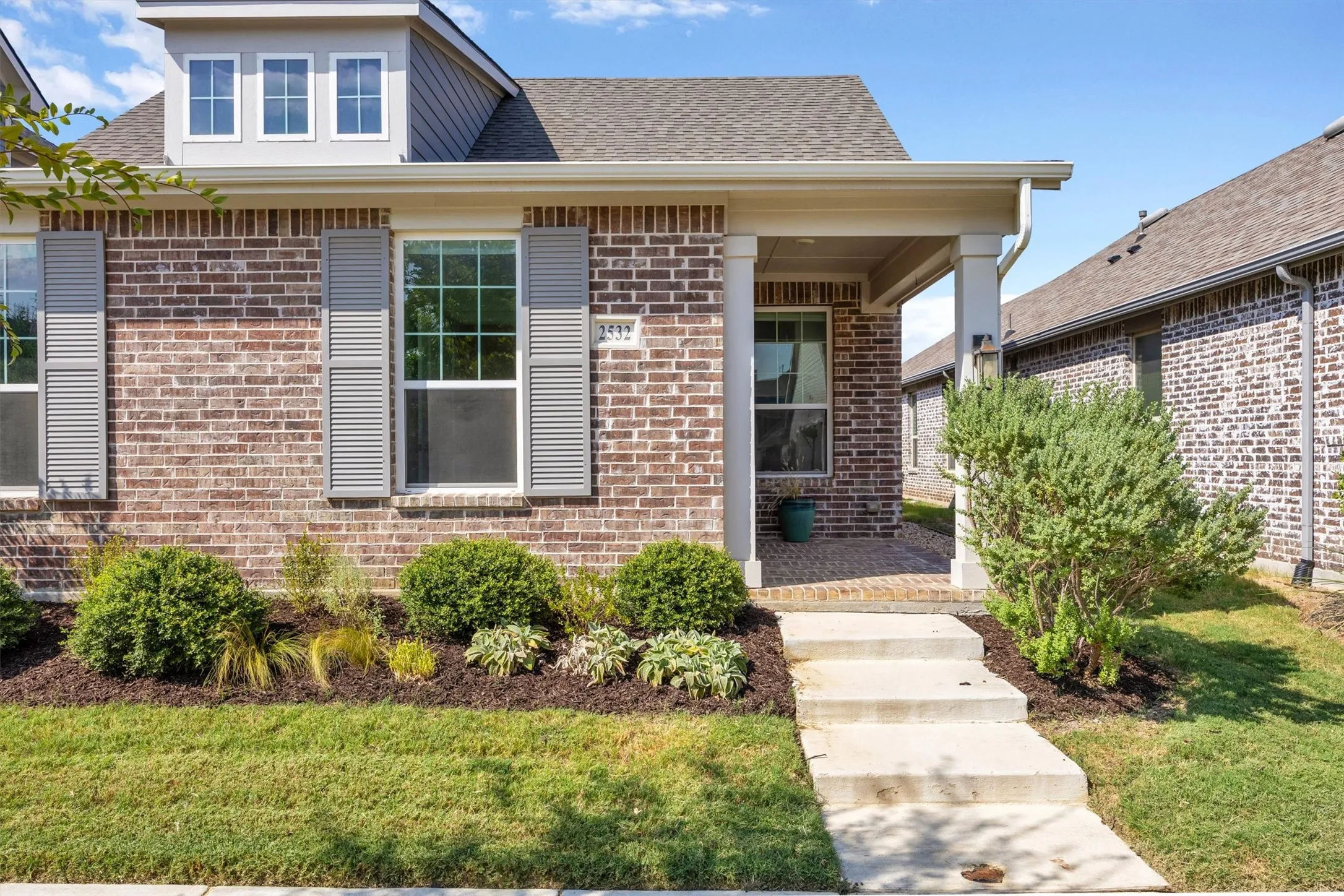 Brick exterior with gray shutters, covered porch, dormer window, and mature landscaping