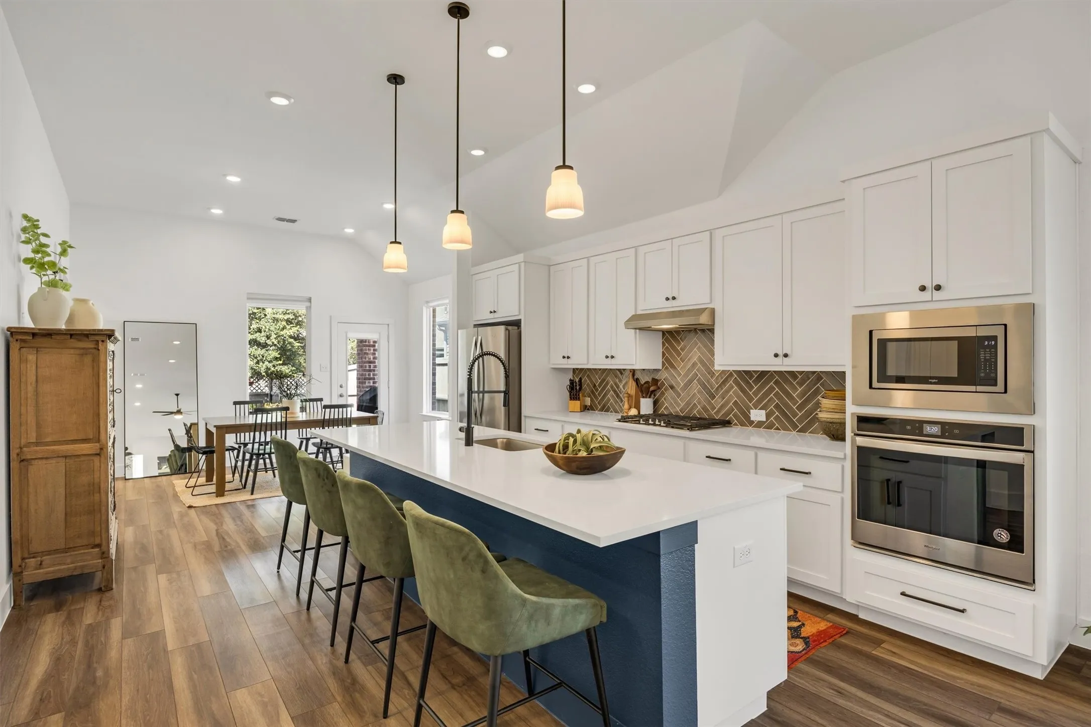 Vaulted ceilings, wood-finish flooring, white cabinetry, stainless steel appliances, and a kitchen island with pendant lighting