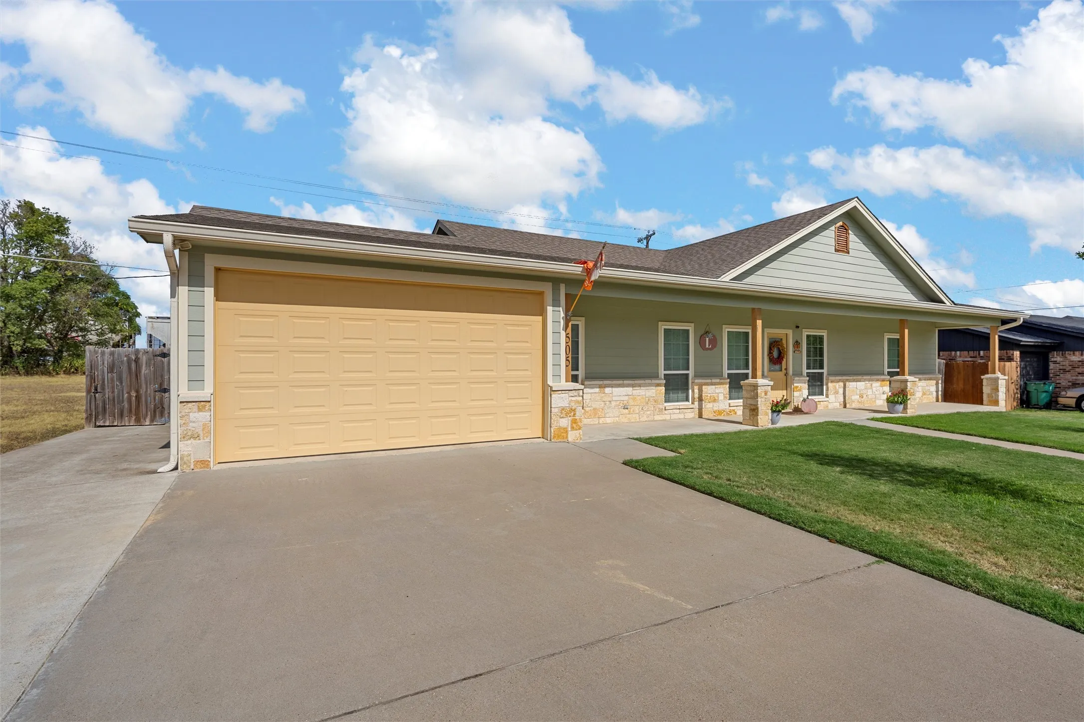 Exterior featuring a light green siding, stone veneer accents, a covered front porch with wood-finish columns, a two-car garage, and a concrete driveway