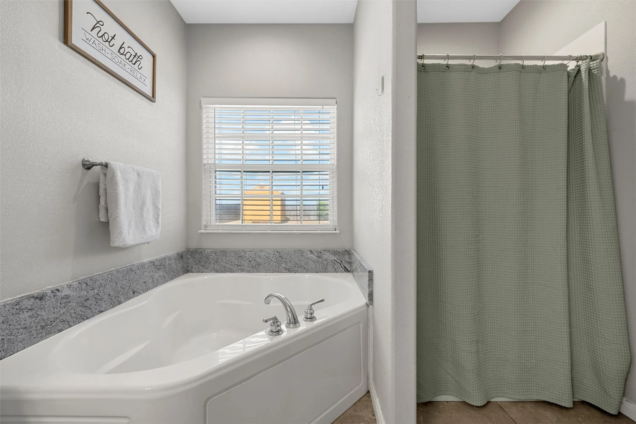 Soaking tub with a polished chrome faucet, stone-finish surround, and a privacy window with blinds
