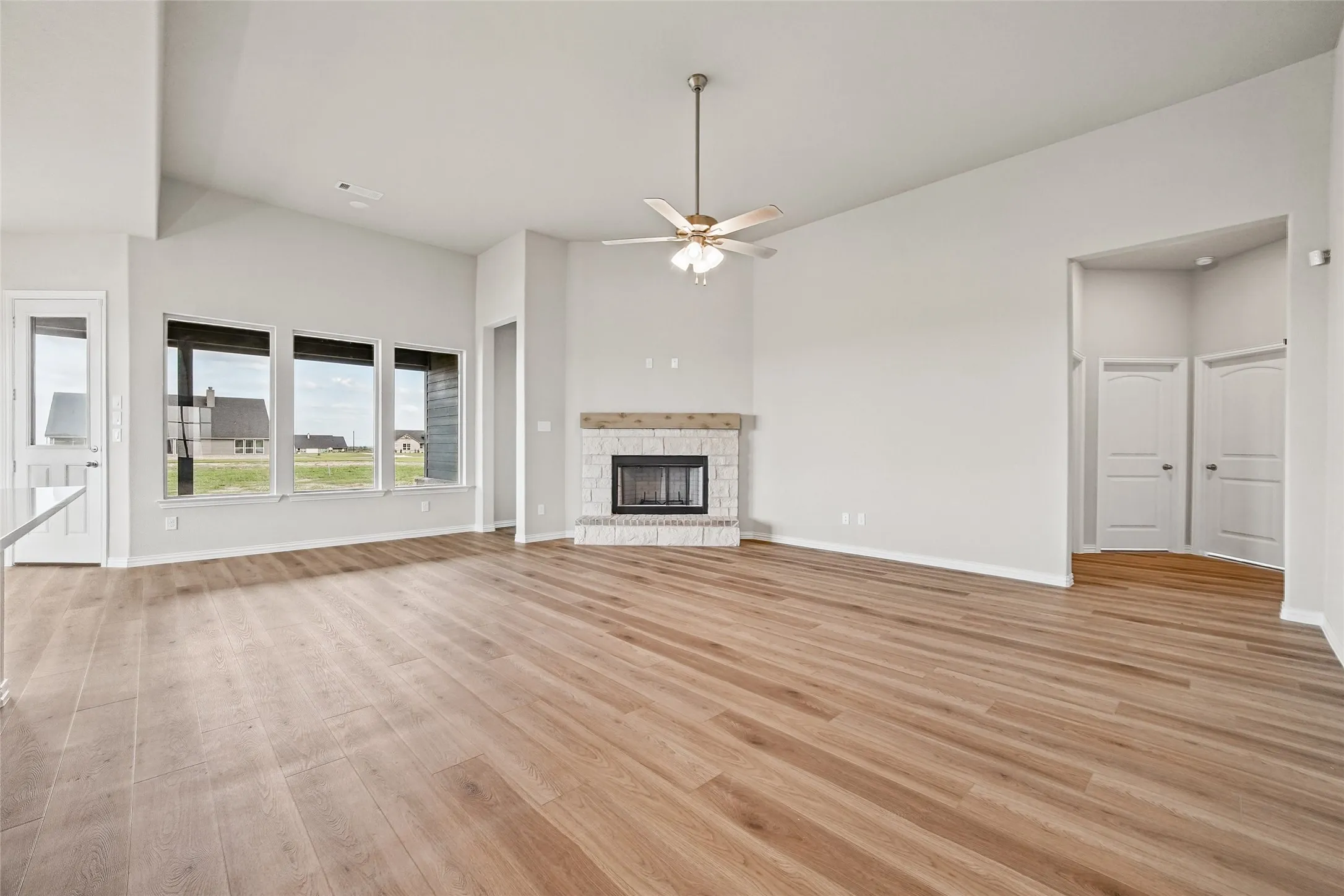 Living area featuring wood-finish flooring, vaulted ceilings, a fireplace with stone detailing and a timber mantel, a multi-blade ceiling fan, and large picture windows