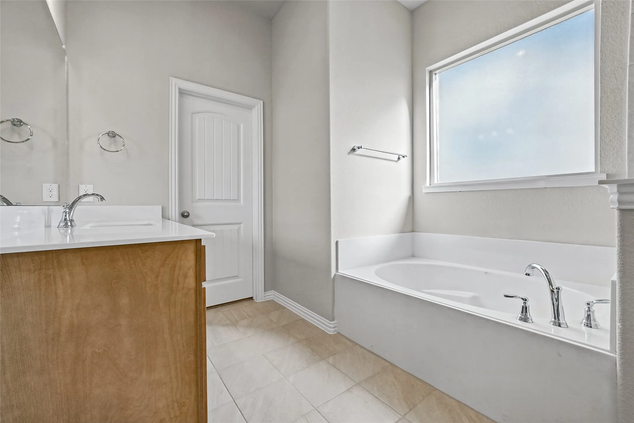 Bathroom featuring a drop-in soaking tub with a deck-mounted chrome faucet, light-toned tile flooring, and a vanity topped with a white surface and chrome fixture