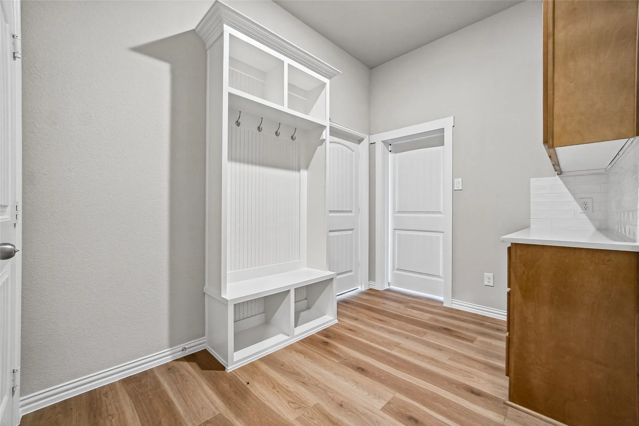 Built-in white mudroom bench with beadboard backing, integrated storage cubbies, and overhead coat hooks, featuring wood-finish flooring, shaker-style cabinetry, and stone-look countertops