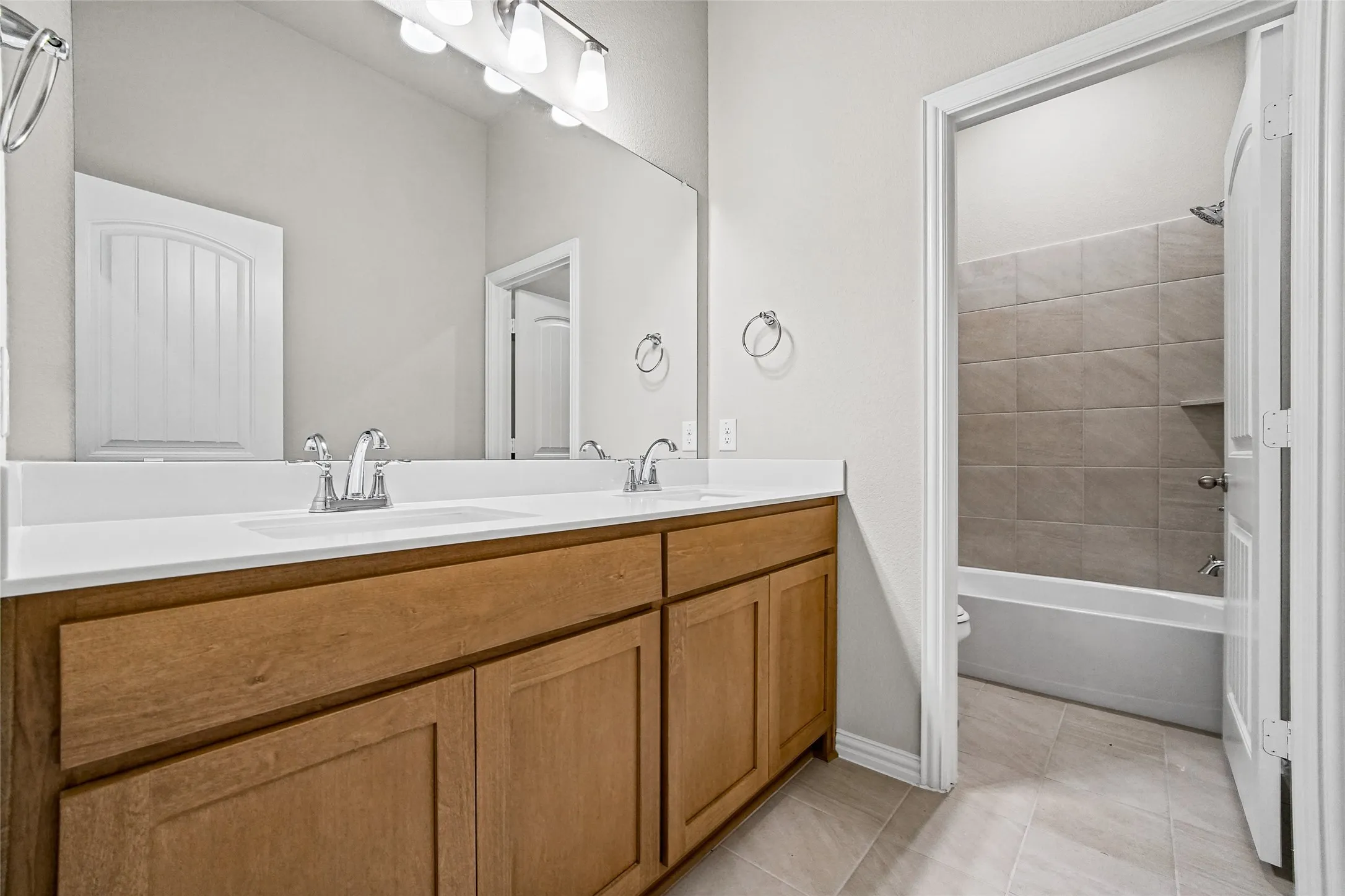 Dual-sink vanity with light wood-finish cabinetry, solid-surface countertop, full-width wall mirror, and chrome finish hardware, with a ceramic-tiled bathtub enclosure