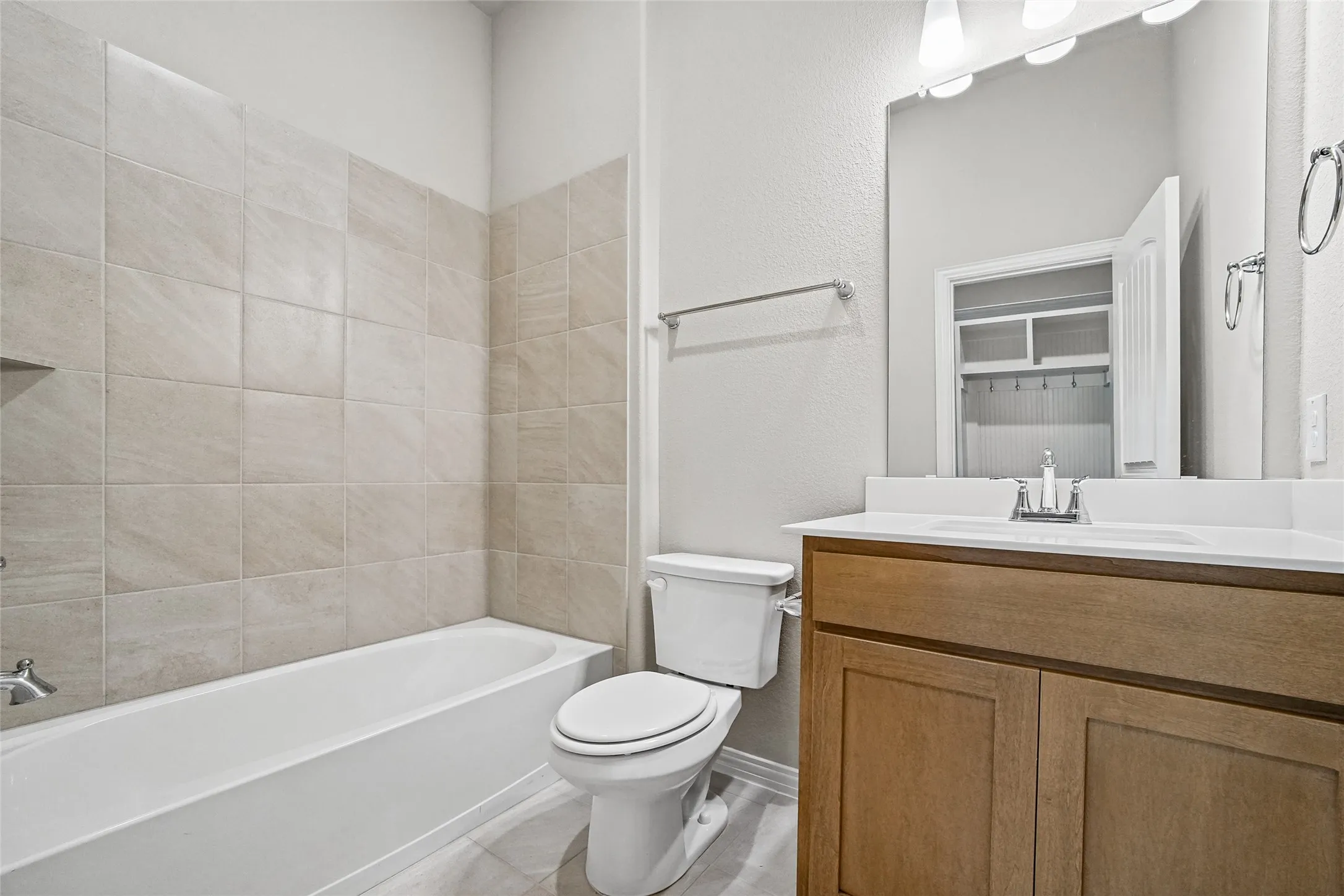 Bathroom featuring a wood-finish vanity with a light-toned solid surface countertop, an integrated porcelain sink, a large wall-mounted mirror, and a shower-tub combination with neutral-toned square tile surround
