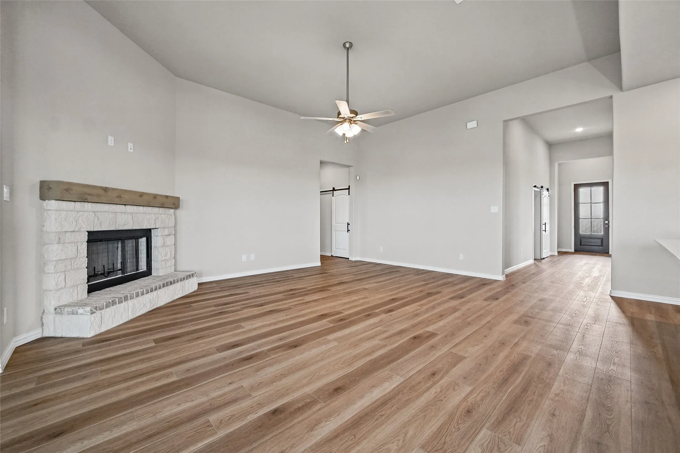High-ceilinged room featuring wood-finish flooring, a stacked-stone fireplace with a wooden mantel, a central ceiling fan, and sliding barn-style doors