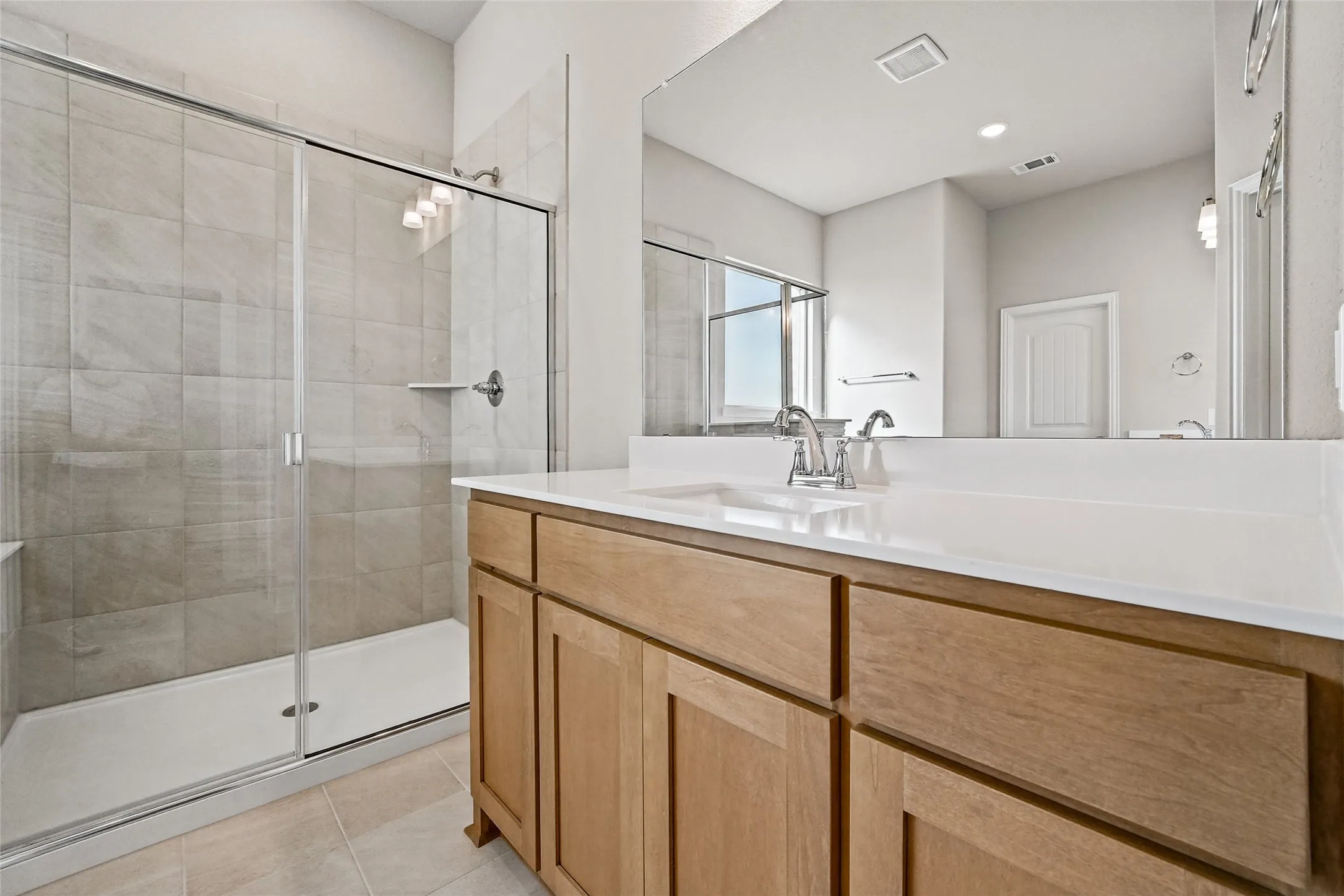 Bathroom featuring a wood-finish vanity with a light-toned countertop, integrated dual sinks, a large wall-mounted mirror, and a walk-in shower with tile surround and glass enclosure