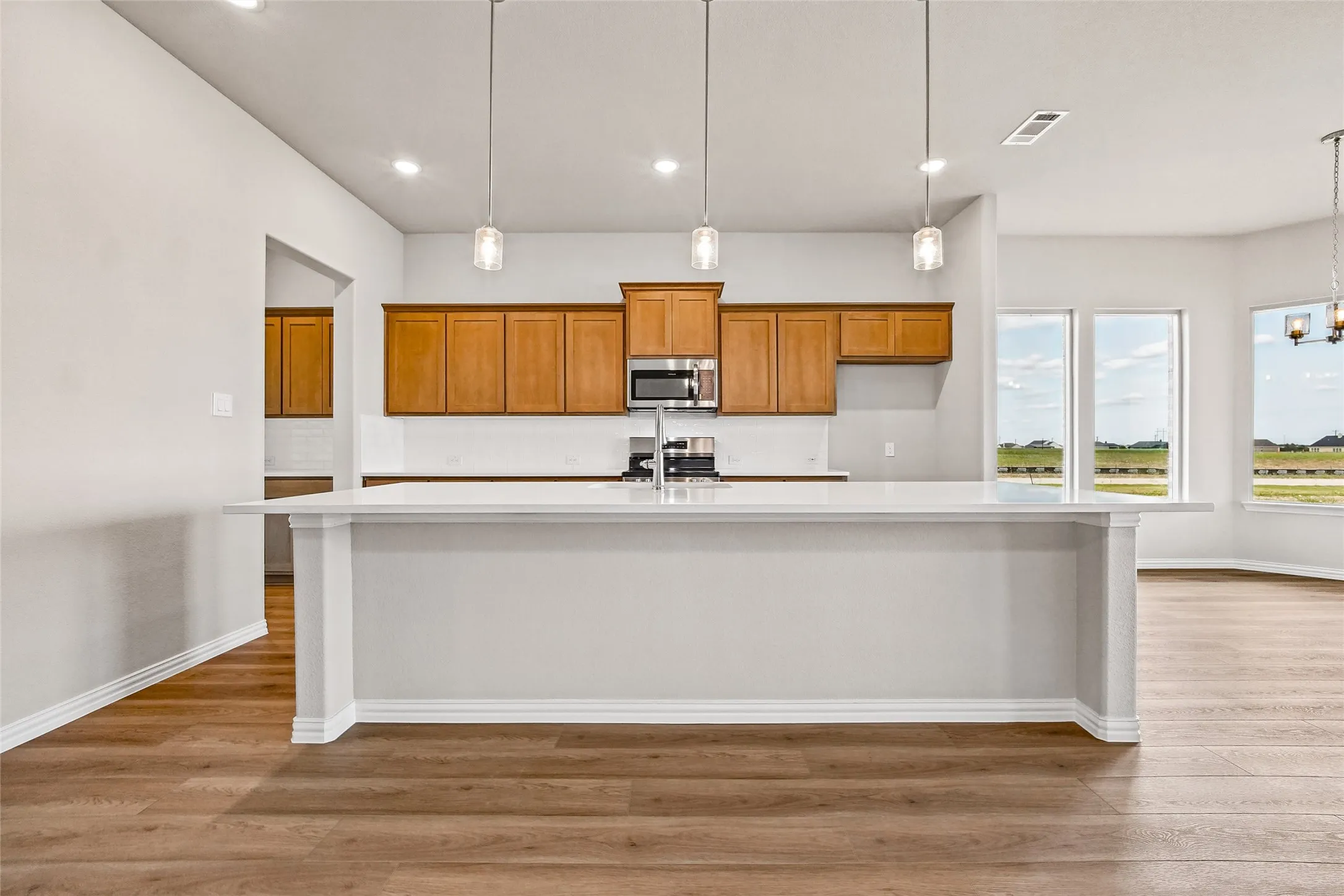 Kitchen featuring an expansive stone-surface island, wood-finish cabinetry, built-in stainless steel microwave, three glass pendant lights, and wood-finish flooring