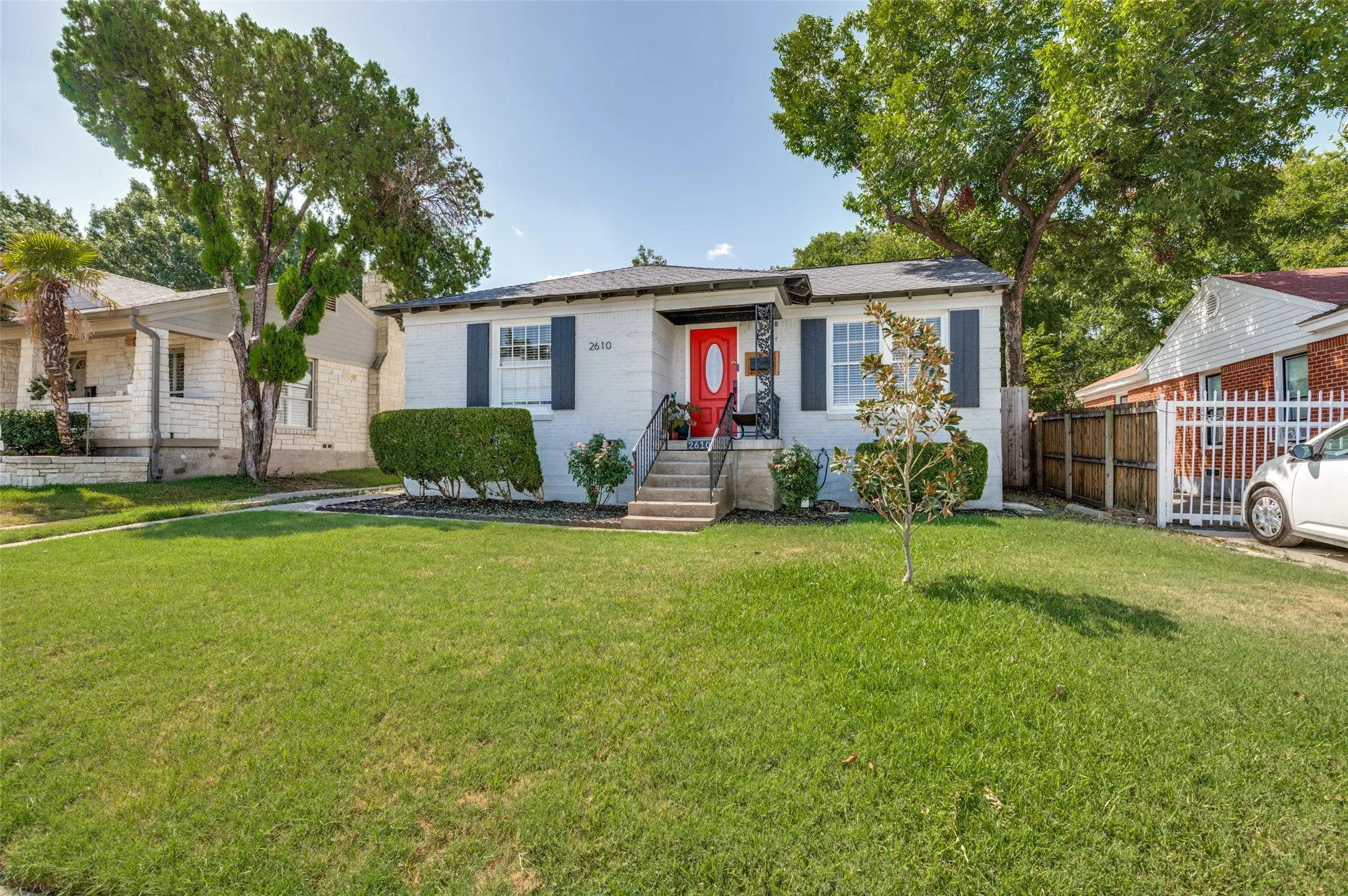 White exterior with dark shutters, red entry door, stone steps with decorative railing, and established front yard landscaping