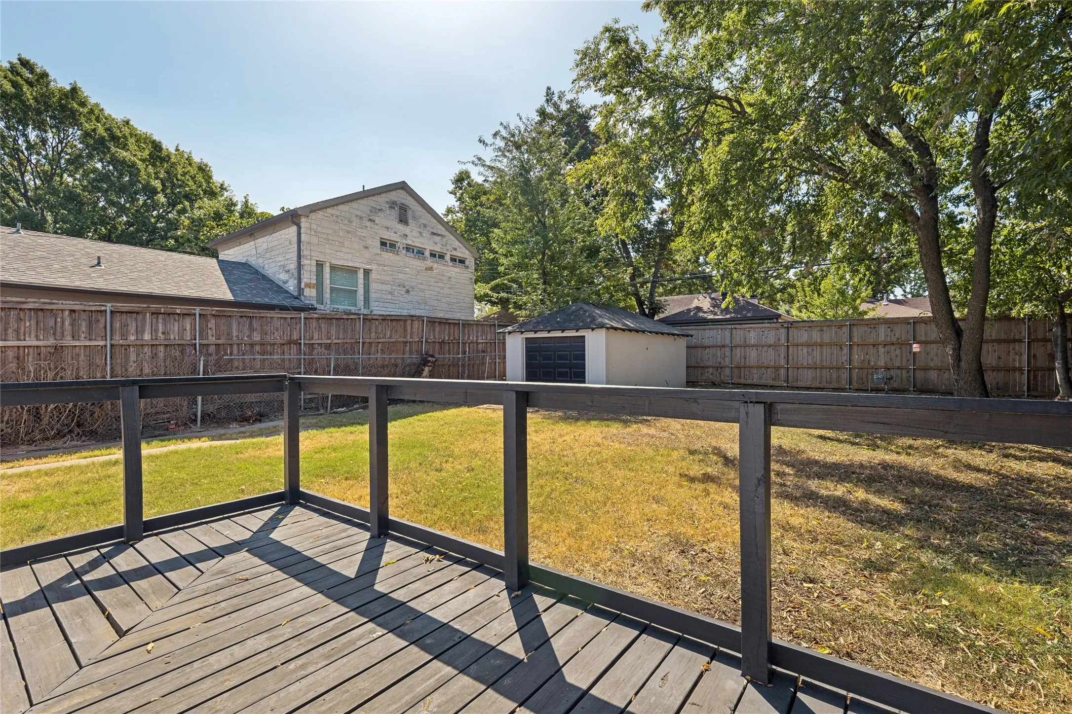 Wood deck with dark railing, expansive lawn, wood privacy fencing, mature trees, and a detached shed