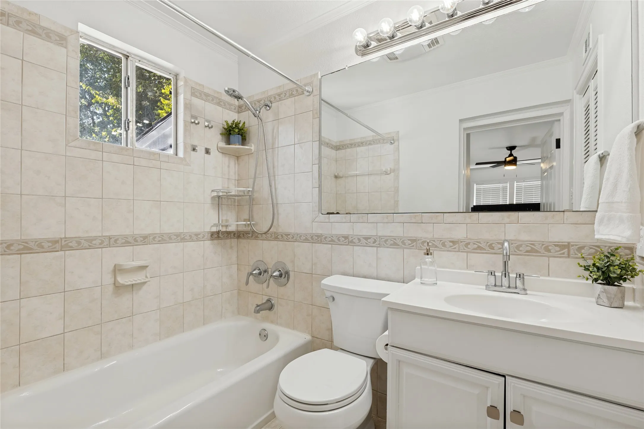 White tiled bathroom featuring a built-in bathtub, a window, a vanity with a white countertop and integrated sink, and a large mirror