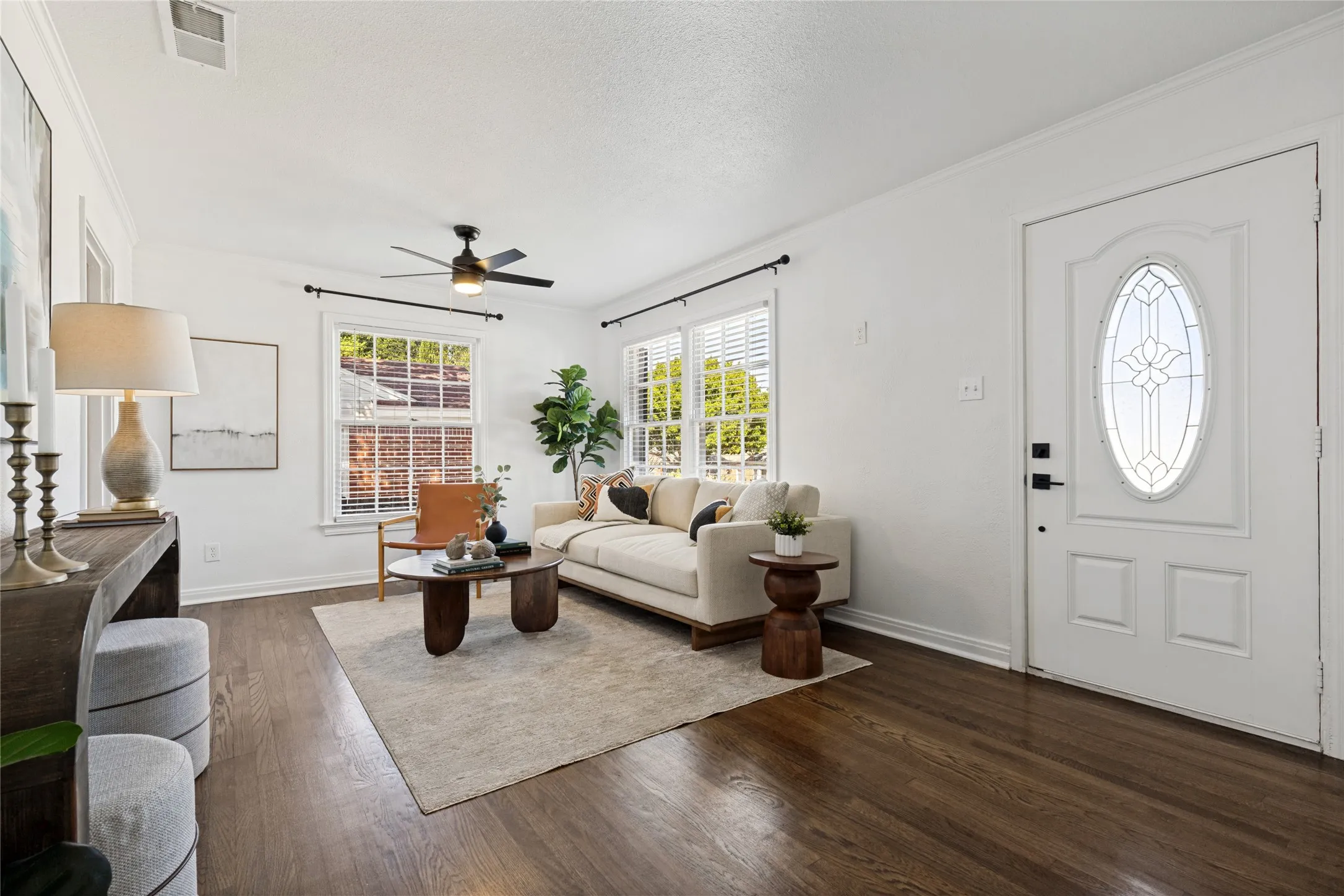 Rich wood-finish flooring, white walls, multiple windows, a ceiling fan, and a front entry door with leaded glass insert