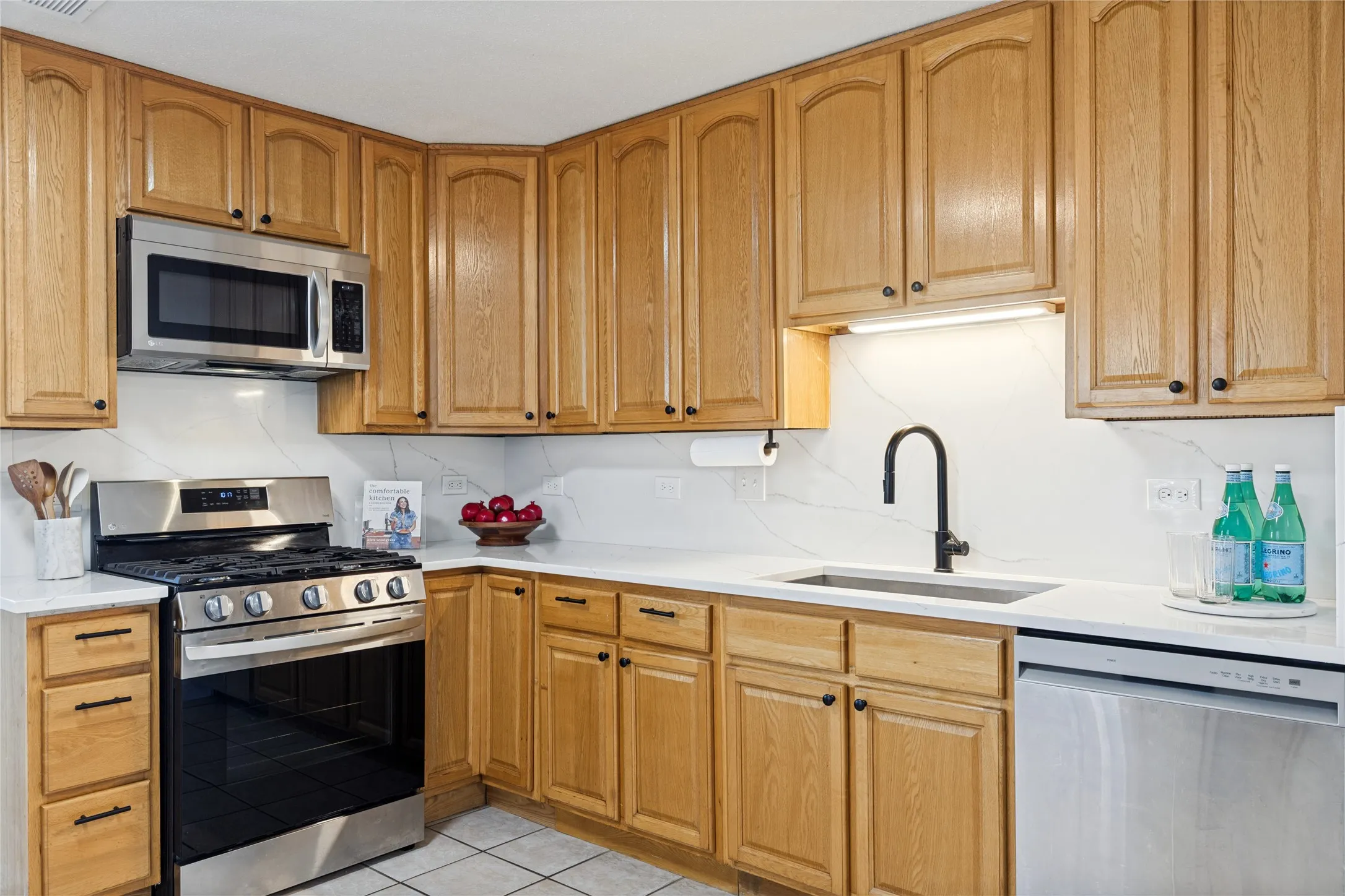 Stainless steel appliances, light wood cabinetry, white countertops, white tile flooring, and a matte black faucet
