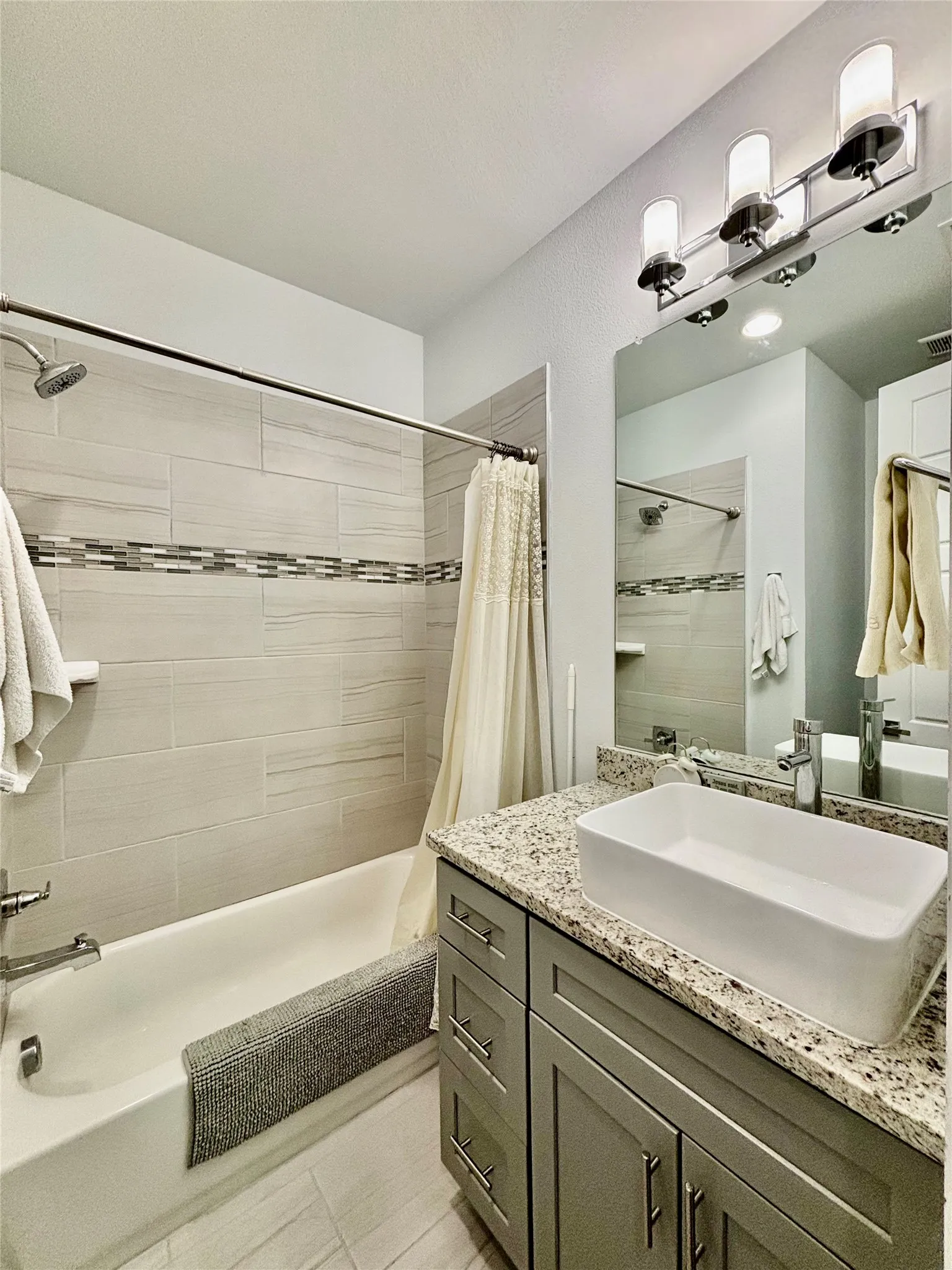 Tub and shower combination with tiled surround, vanity with stone-finish countertop, rectangular vessel sink, and mounted light fixture
