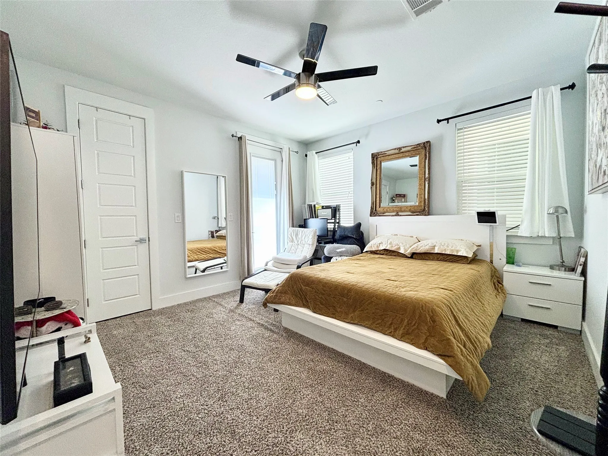 Plush carpeted flooring, modern ceiling fan with light, white paneled door, and multiple windows with blinds