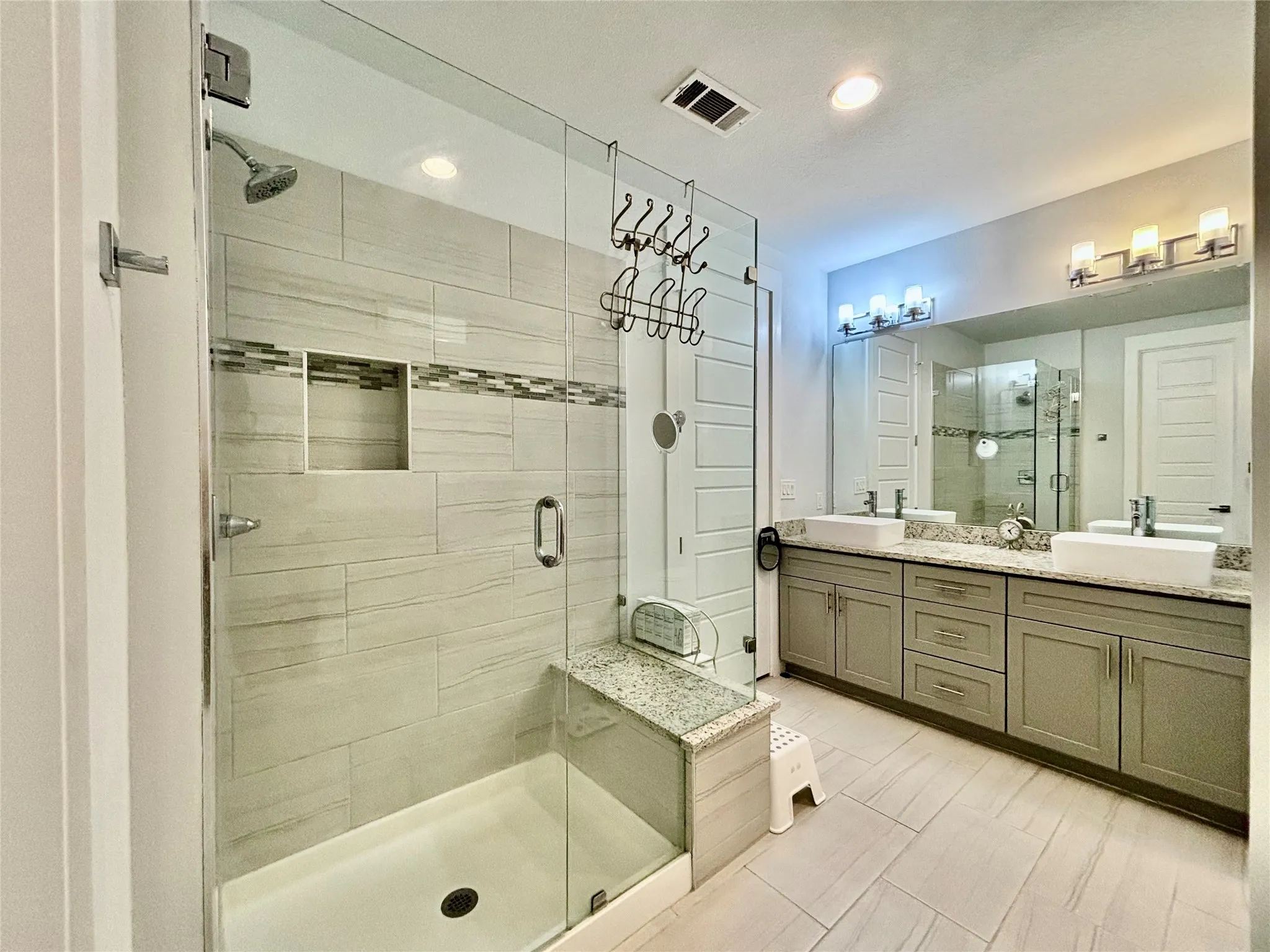 Glass-enclosed shower with a built-in bench and recessed niche, dual vessel sinks, extensive vanity with under-cabinet storage, and tile flooring