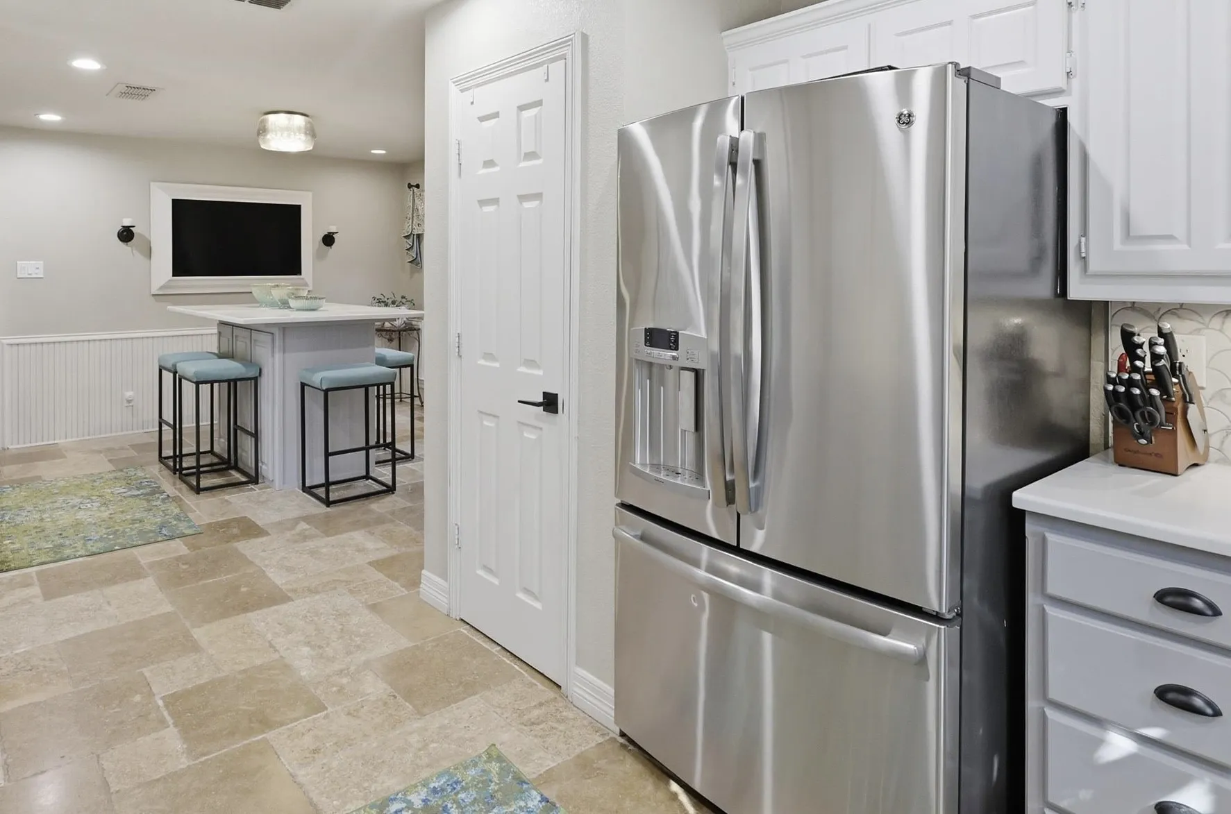 Stainless steel refrigerator, white cabinetry, light countertops, neutral tone tile flooring, and wainscoting