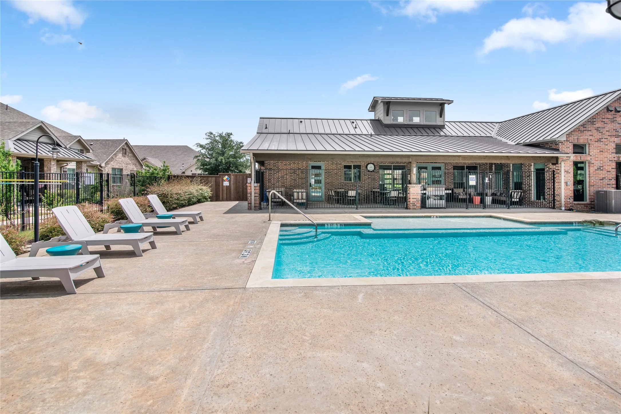 Resort-style resort pool with crystal-clear waters and sun loungers, anchored by a modern clubhouse featuring metal roofing, brick construction, and expansive glass doors