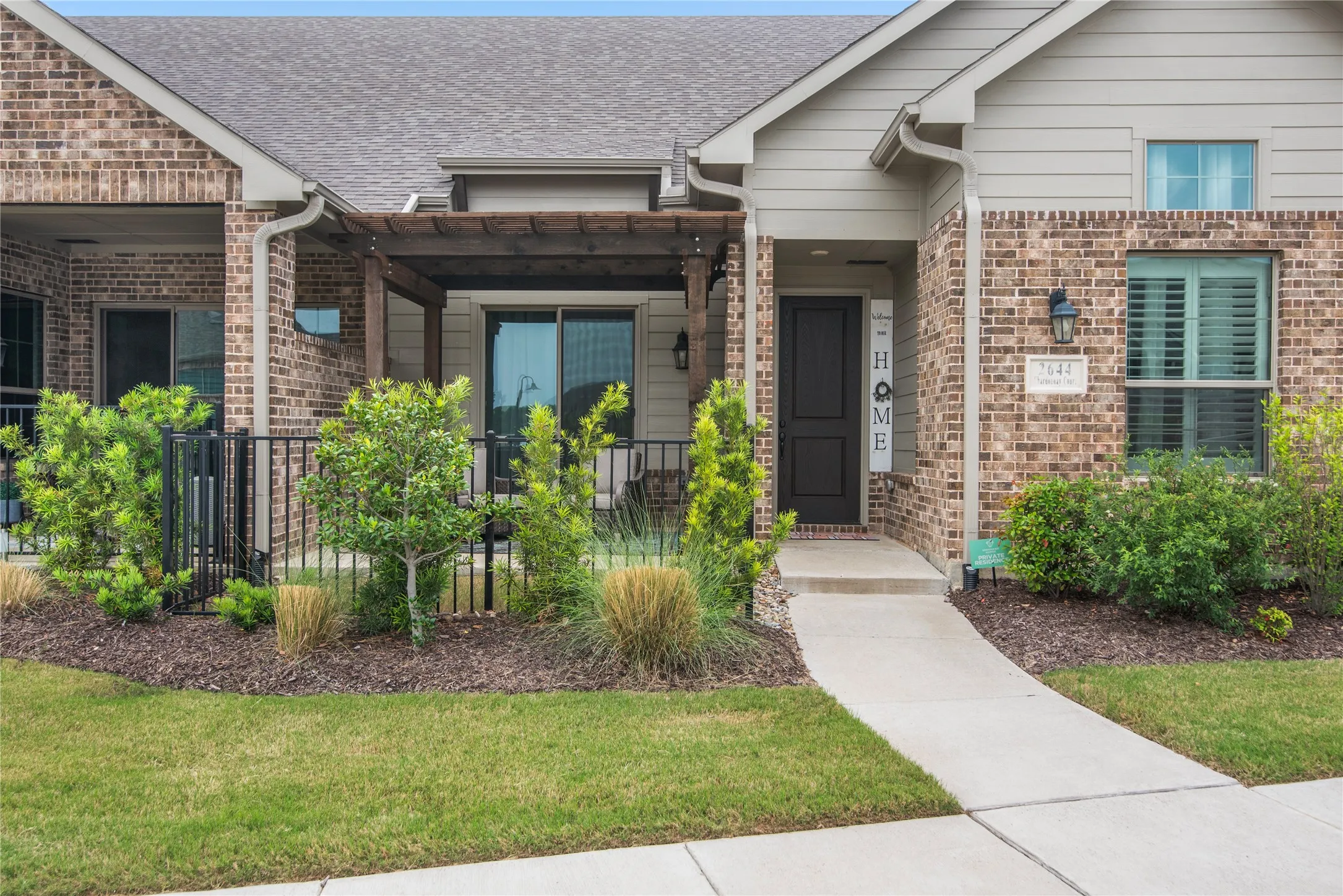 Brick and siding exterior, front entry with dark paneled door, covered patio with wood pergola, mature landscaping, and concrete walkway