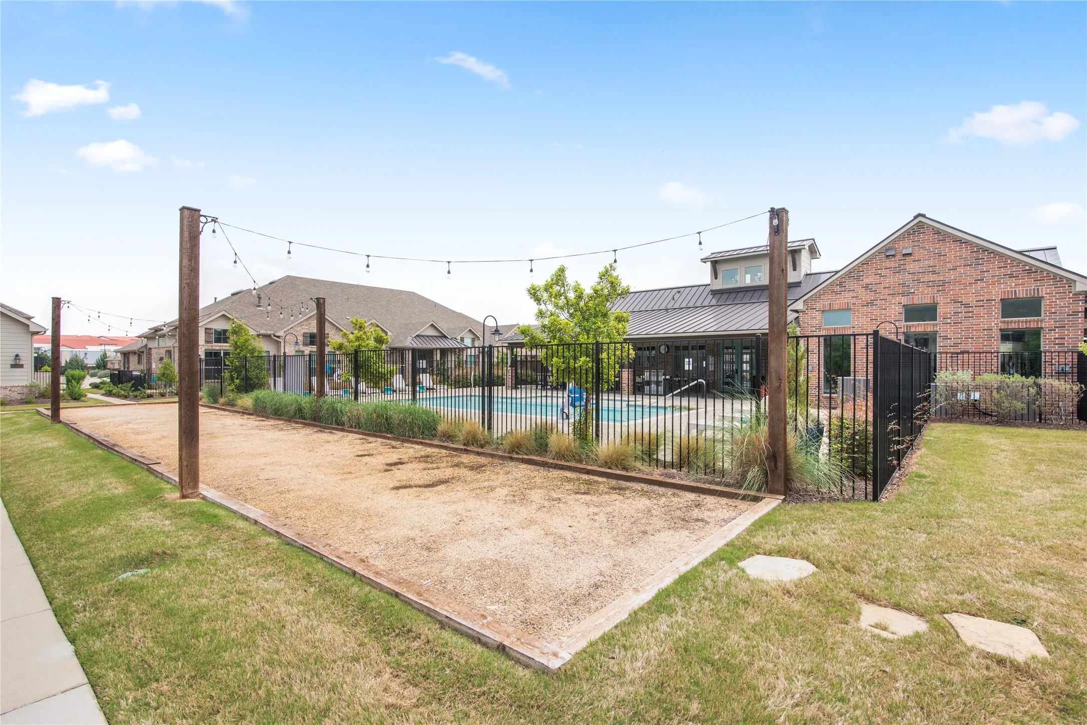 Bocce ball court with string lighting, adjacent to a fenced swimming pool and brick structure