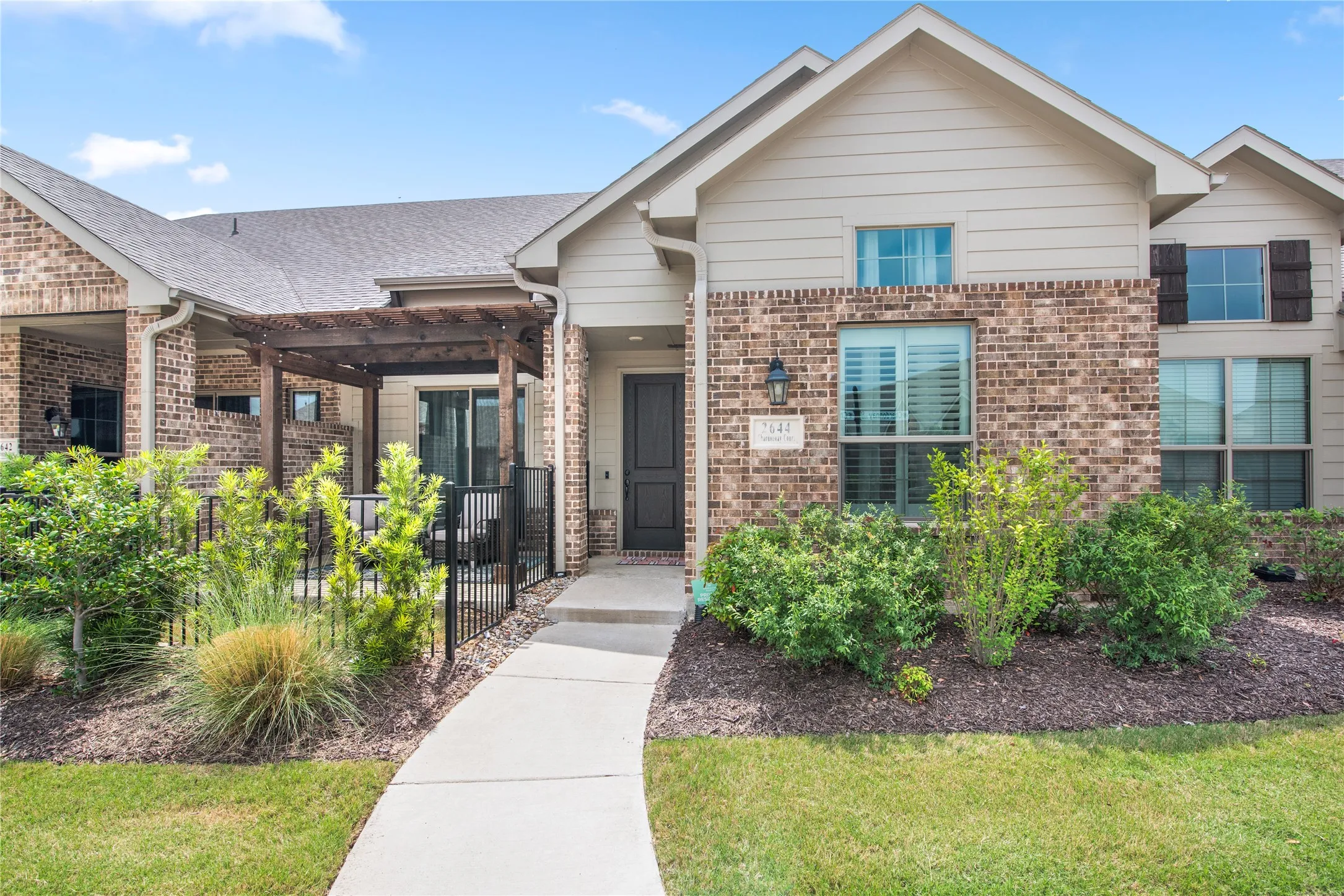 Brick and siding exterior with a concrete walkway, mature landscaping, and a wood-finish pergola