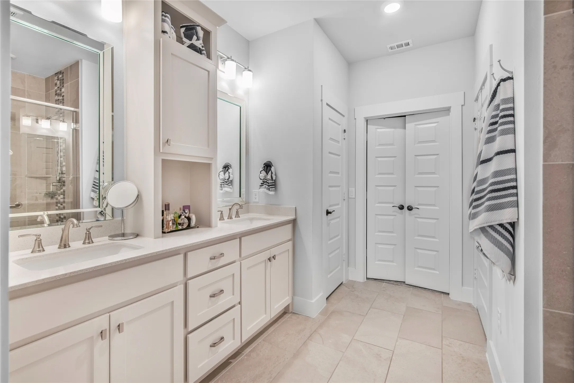 Dual vanity with undermount sinks, white cabinetry, integrated storage tower, light tile flooring, and a tiled shower with a glass enclosure