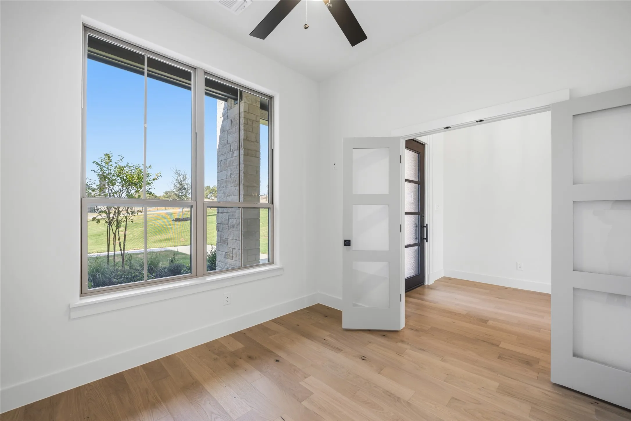 Hardwood-finish flooring, a large window with exterior stone accents, white walls, and a ceiling fan