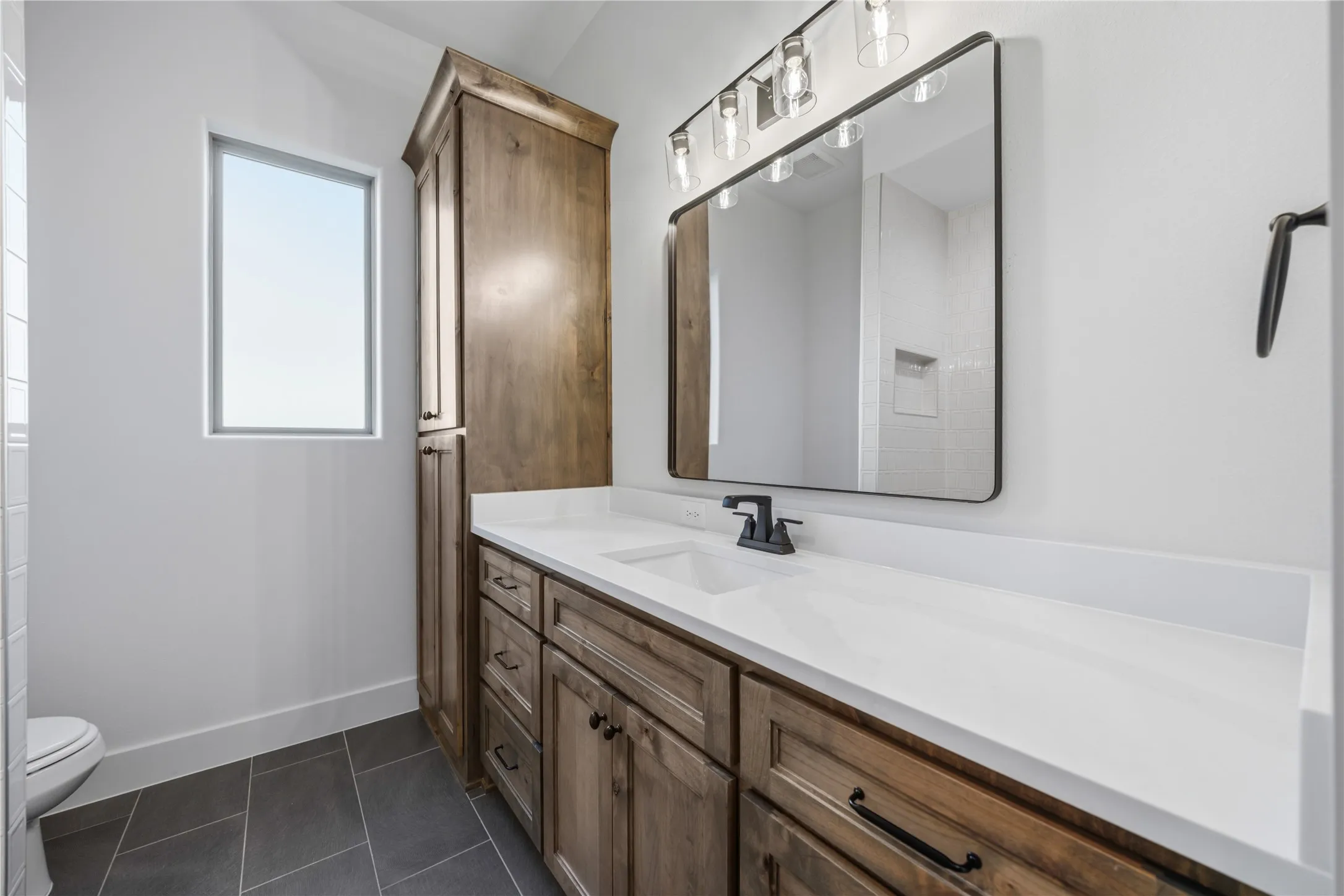 Spacious vanity with a white countertop and integrated sink, dark wood-finish cabinetry with black hardware, and an overhead vanity light fixture