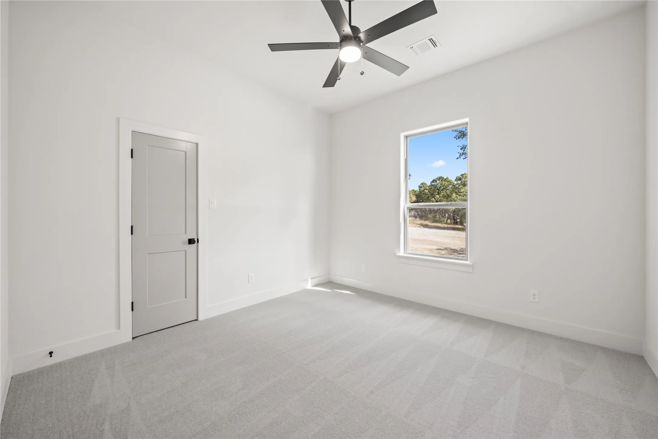 Light gray carpet flooring, white painted walls, single window with outdoor scenery, ceiling fan with integrated lighting, and a two-panel interior door with matte black hardware
