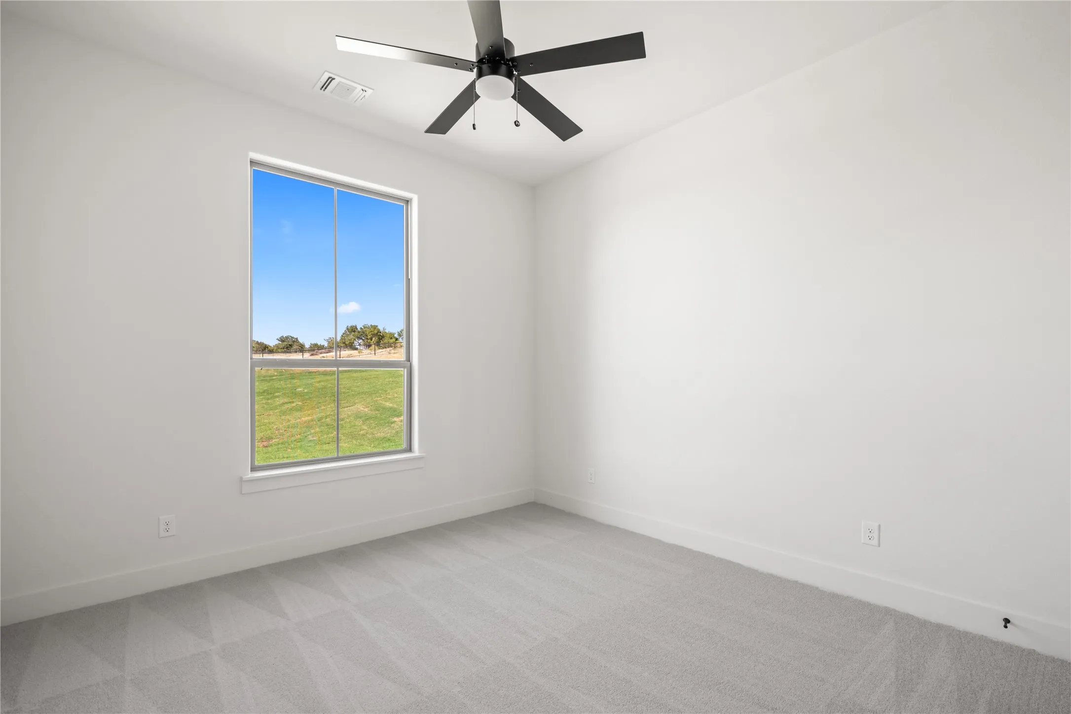 Light gray walls with a large window overlooking a field, complemented by a black ceiling fan and gray patterned carpeting