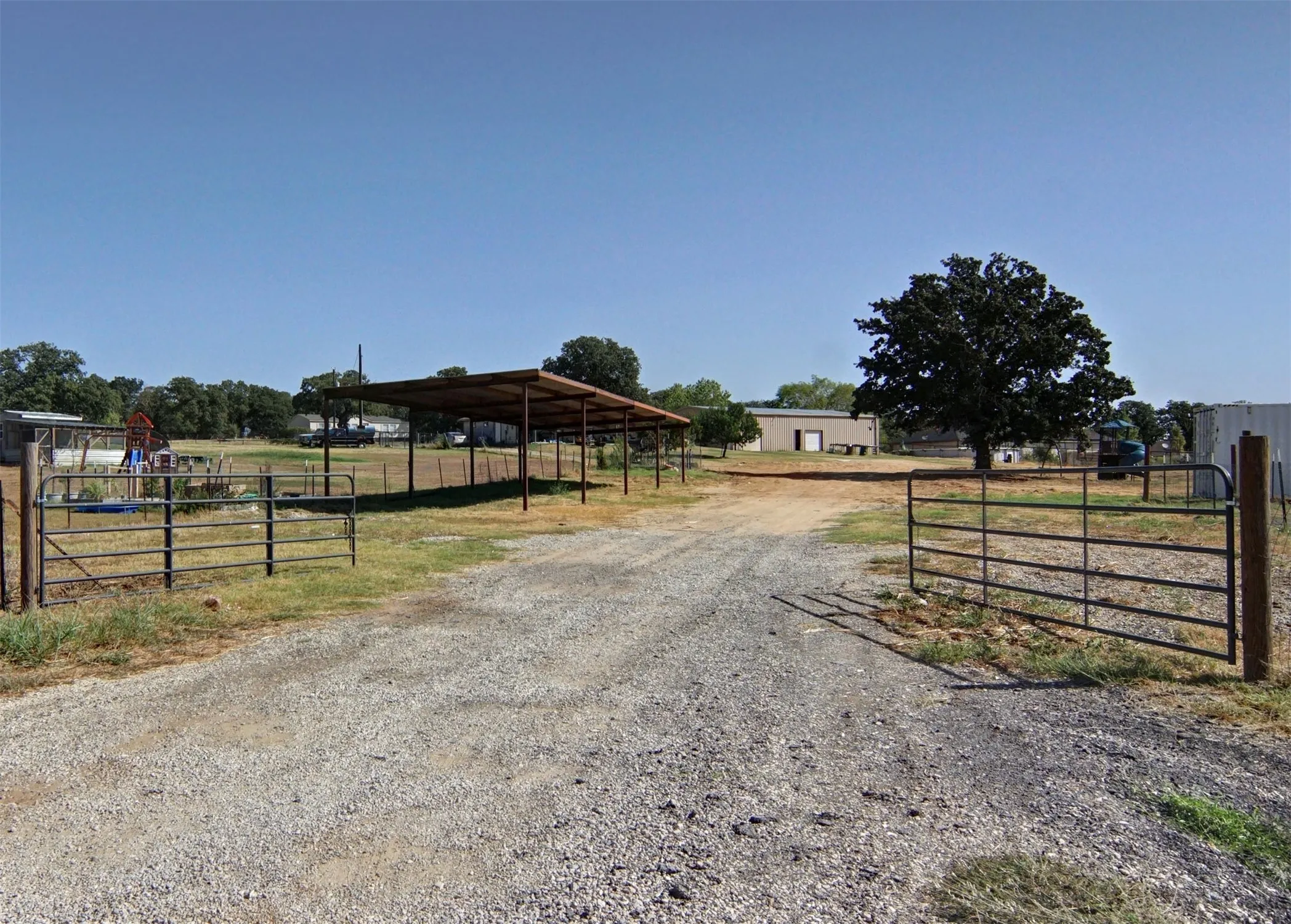 Gravel driveway with metal gates leading to an open-air structure and a mature tree