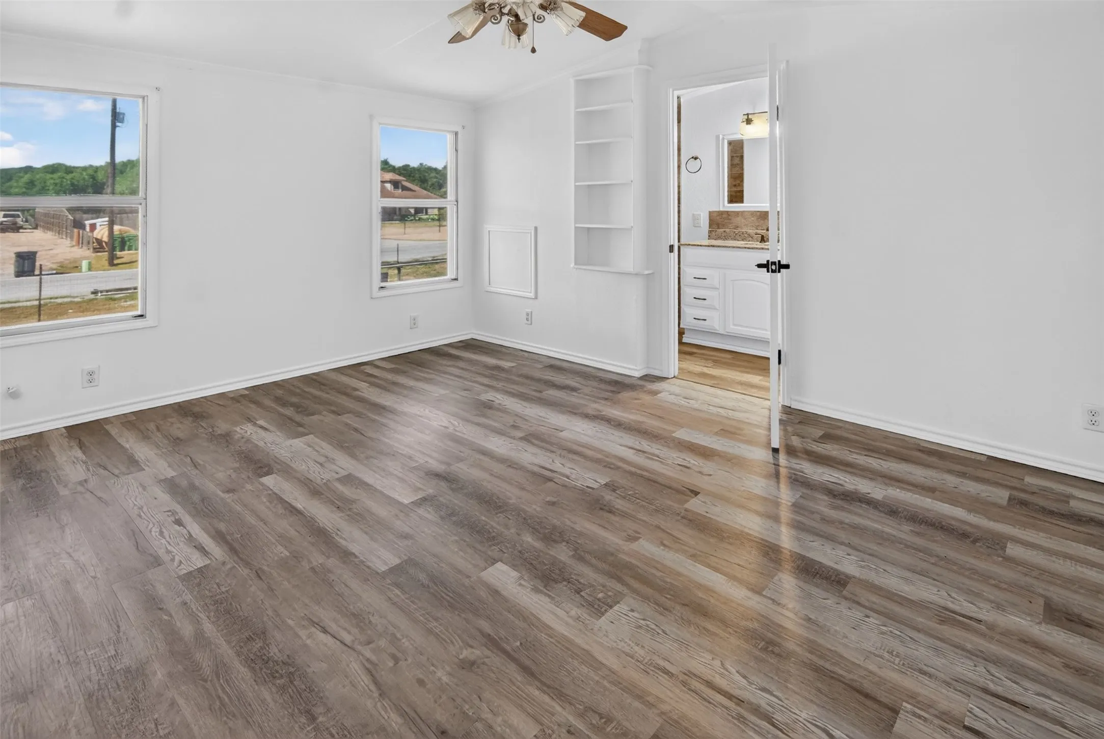 Wood-finish flooring, white walls, built-in shelving, ceiling fan, and an en-suite bathroom with white cabinetry