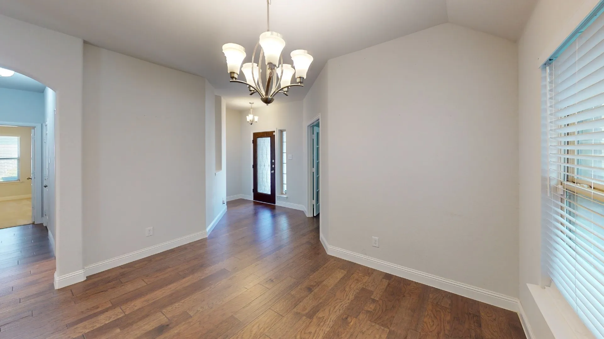 Vaulted ceiling with a multi-arm chandelier, wood-finish flooring, an arched doorway, and a window with blinds
