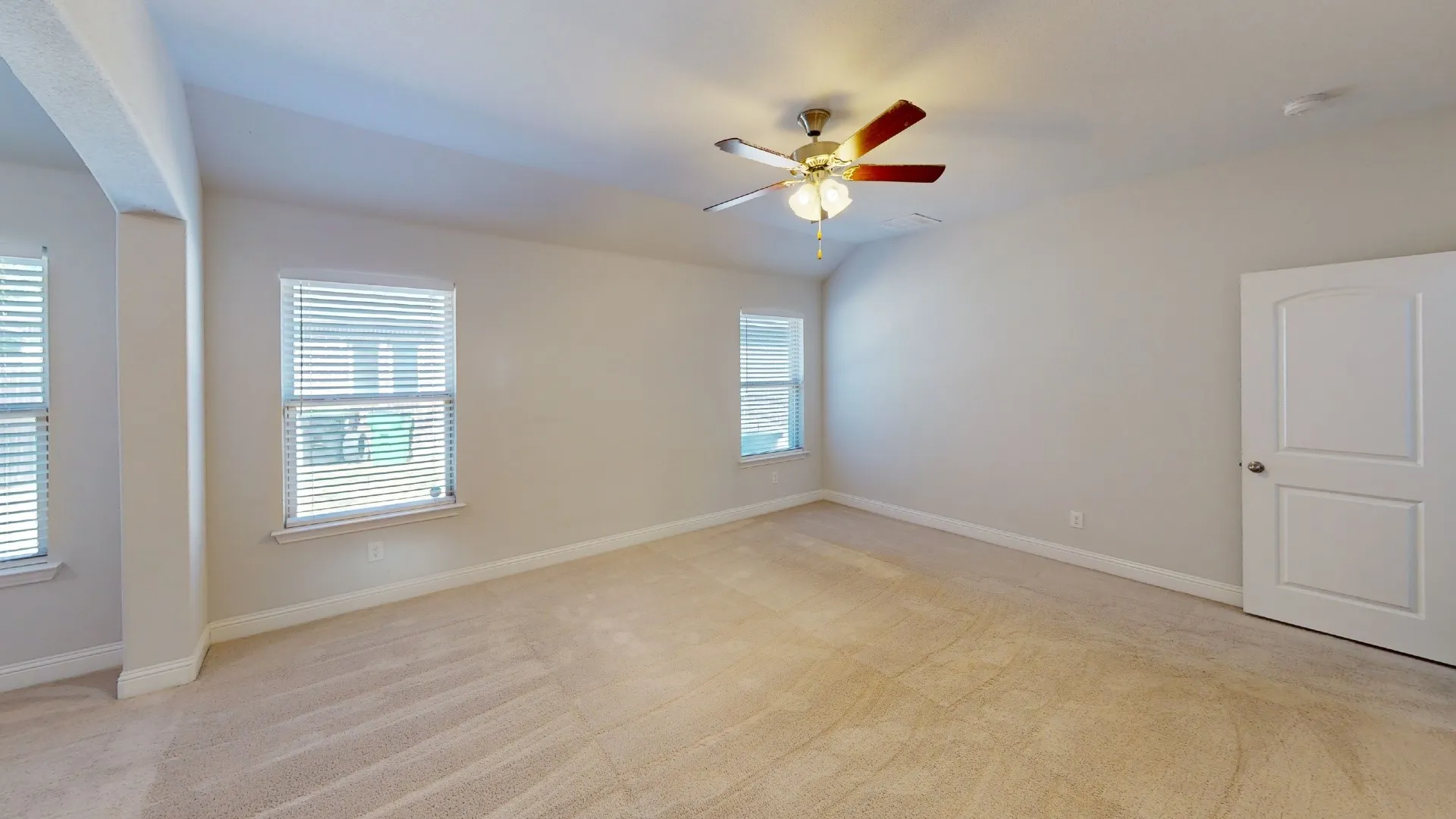 Spacious room with plush carpeting, multiple windows with blinds, a ceiling fan with integrated lighting, and a paneled door