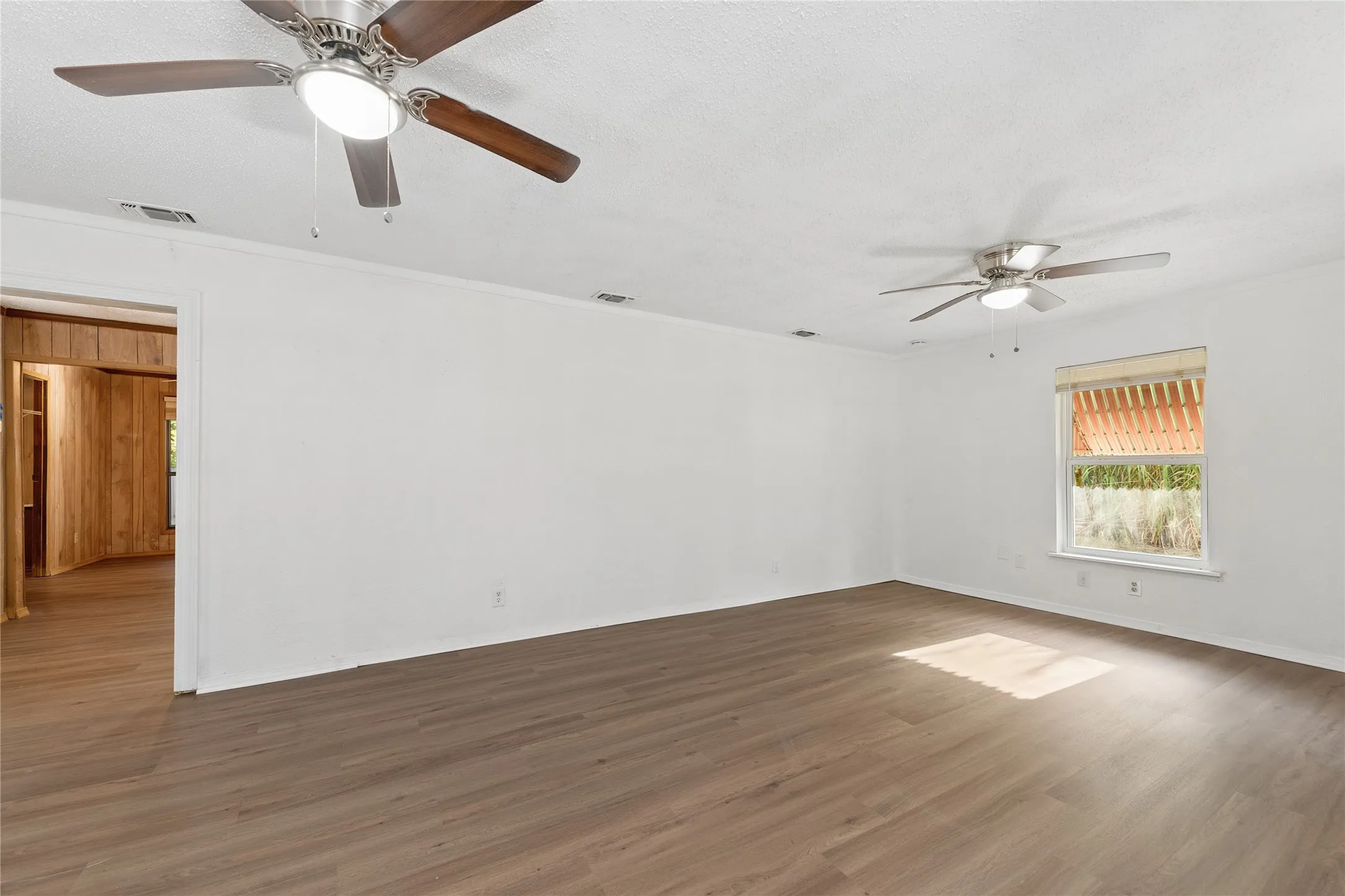 Spacious room featuring wood-finish flooring, two ceiling fans with integrated lighting, and a window with blinds