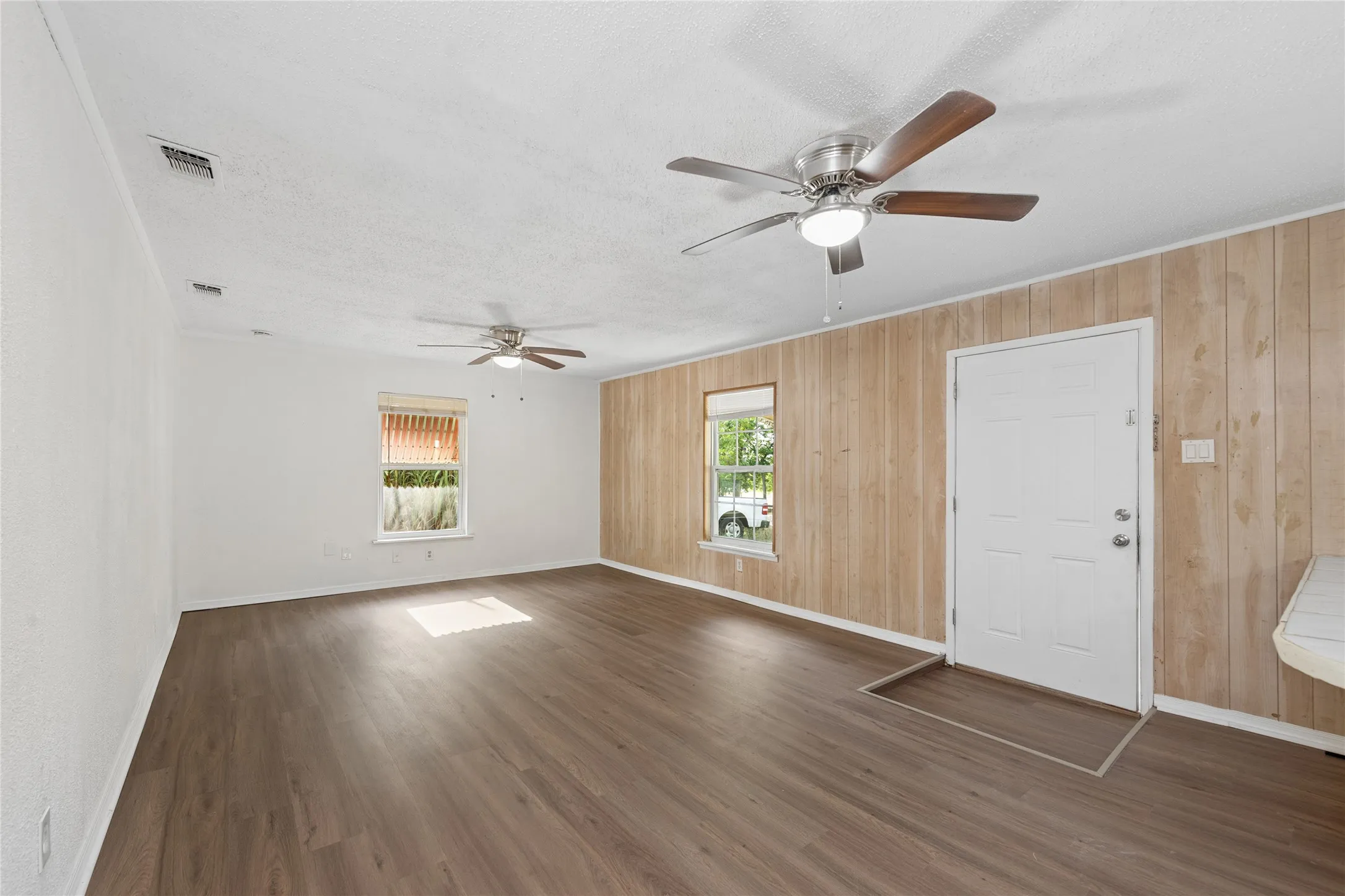 Spacious room featuring wood-finish flooring, two windows, and two ceiling fans with integrated lighting