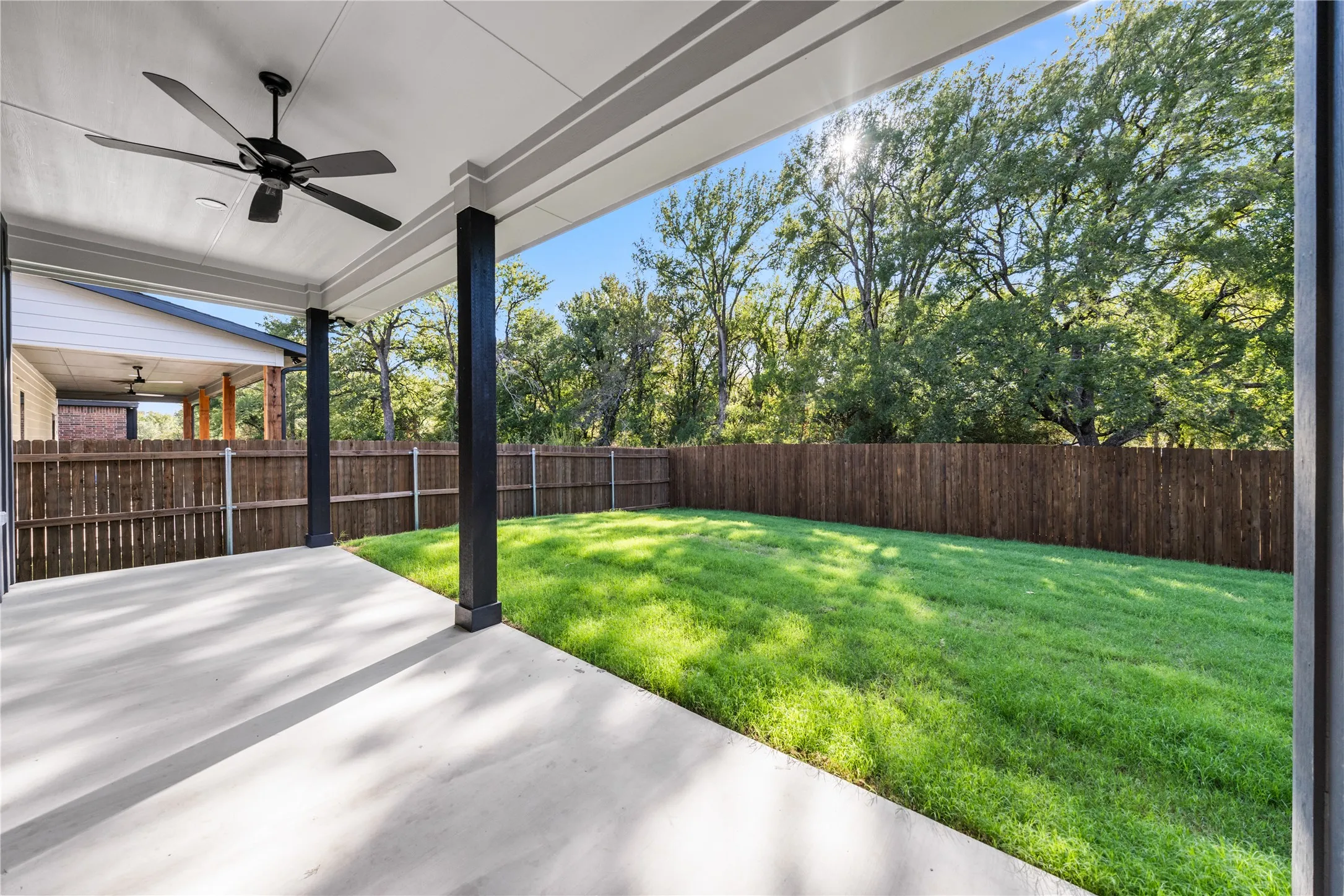 Covered patio with an attached ceiling fan, concrete flooring, and a fenced yard with mature trees