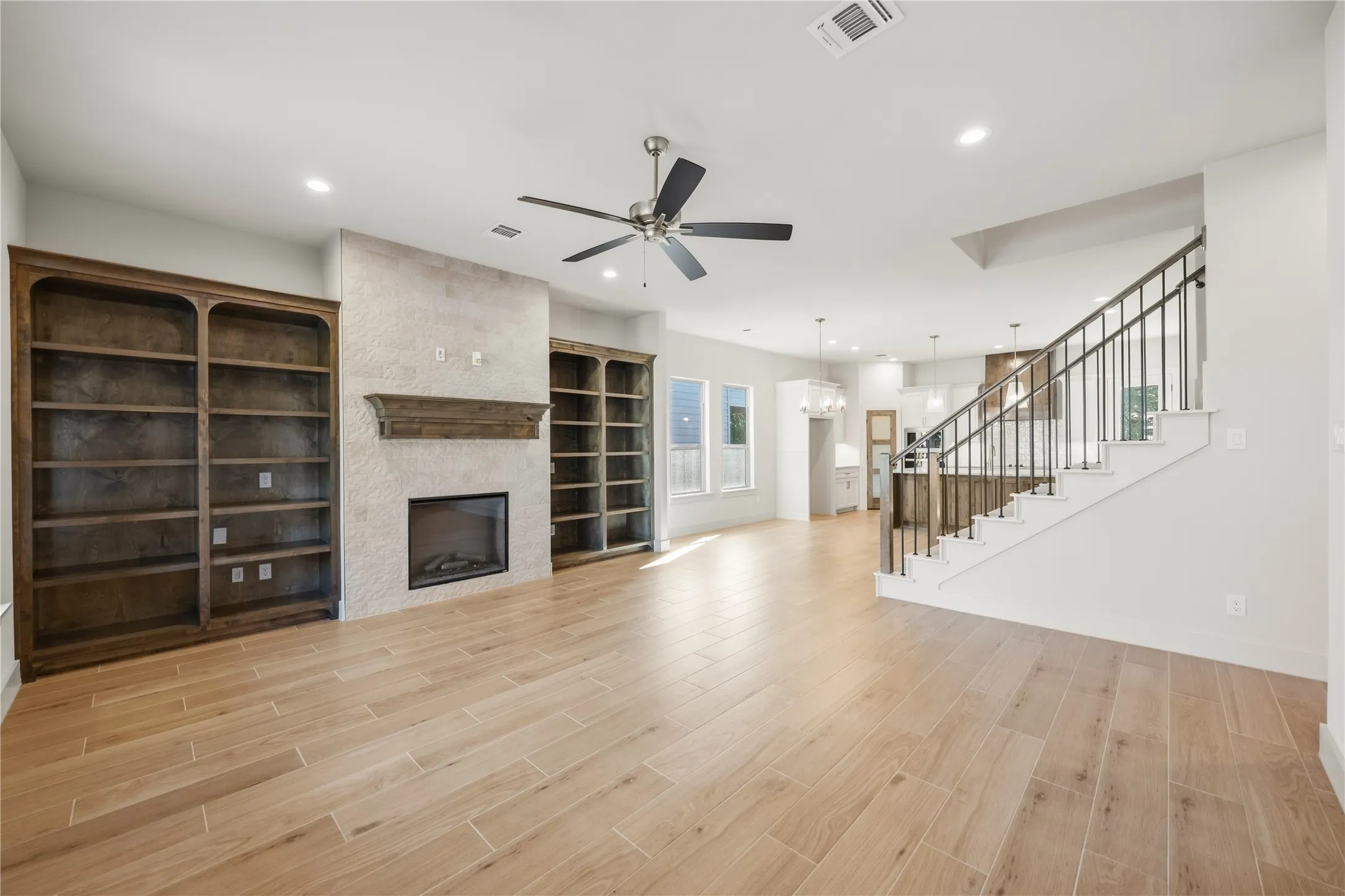 Wood-finish flooring, built-in shelving, fireplace with mantel, ceiling fan, and staircase with metal​​‌​​​​‌​​‌‌​‌‌​​​‌‌​‌​‌​‌​​​‌​​ balusters