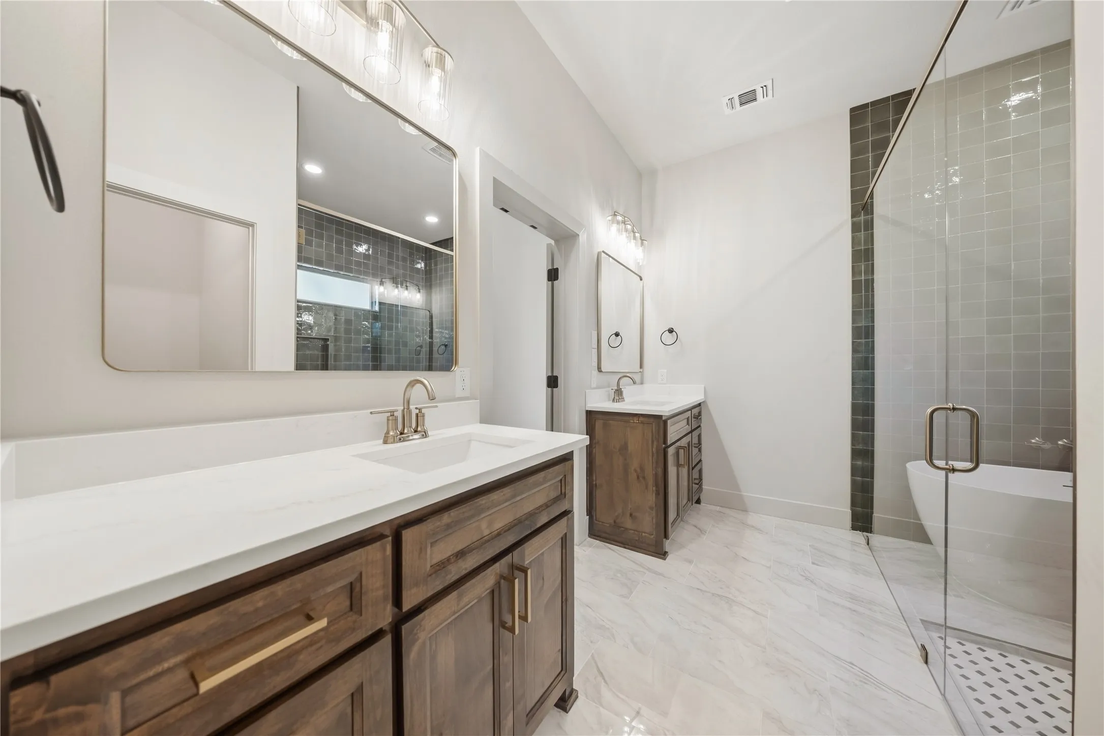 Dual wood-finish vanities with white countertops, glass-enclosed shower, freestanding tub, and large format floor tiling
