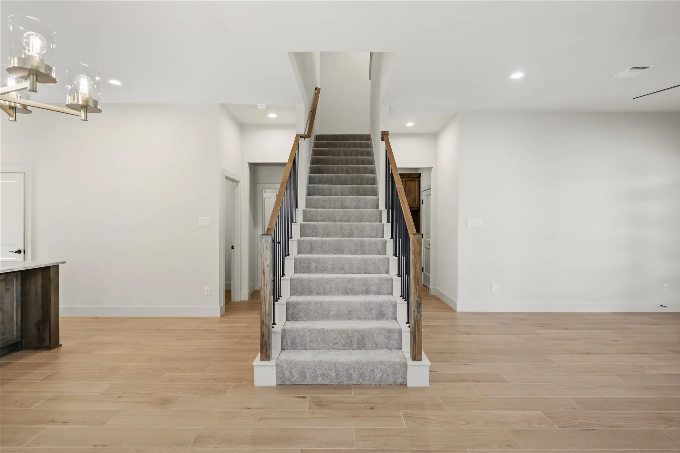Light wood-finish flooring, staircase with light gray carpeting, dark wood-tone handrail, black metal balusters, and recessed ceiling lighting