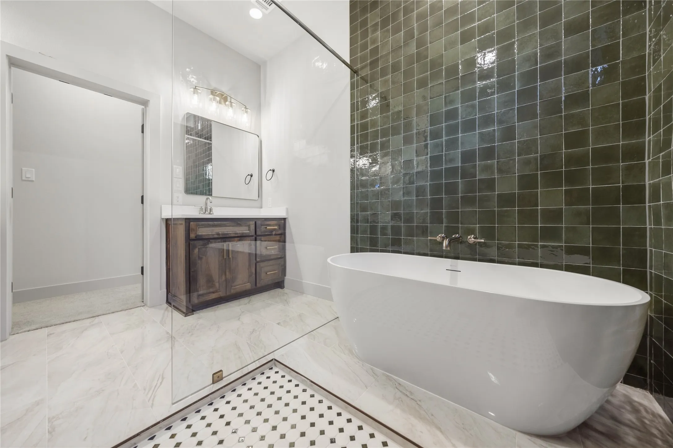 Freestanding soaking tub, floor-to-ceiling square tile wall, glass-enclosed shower, wood-finish vanity with countertop, and a vanity light fixture