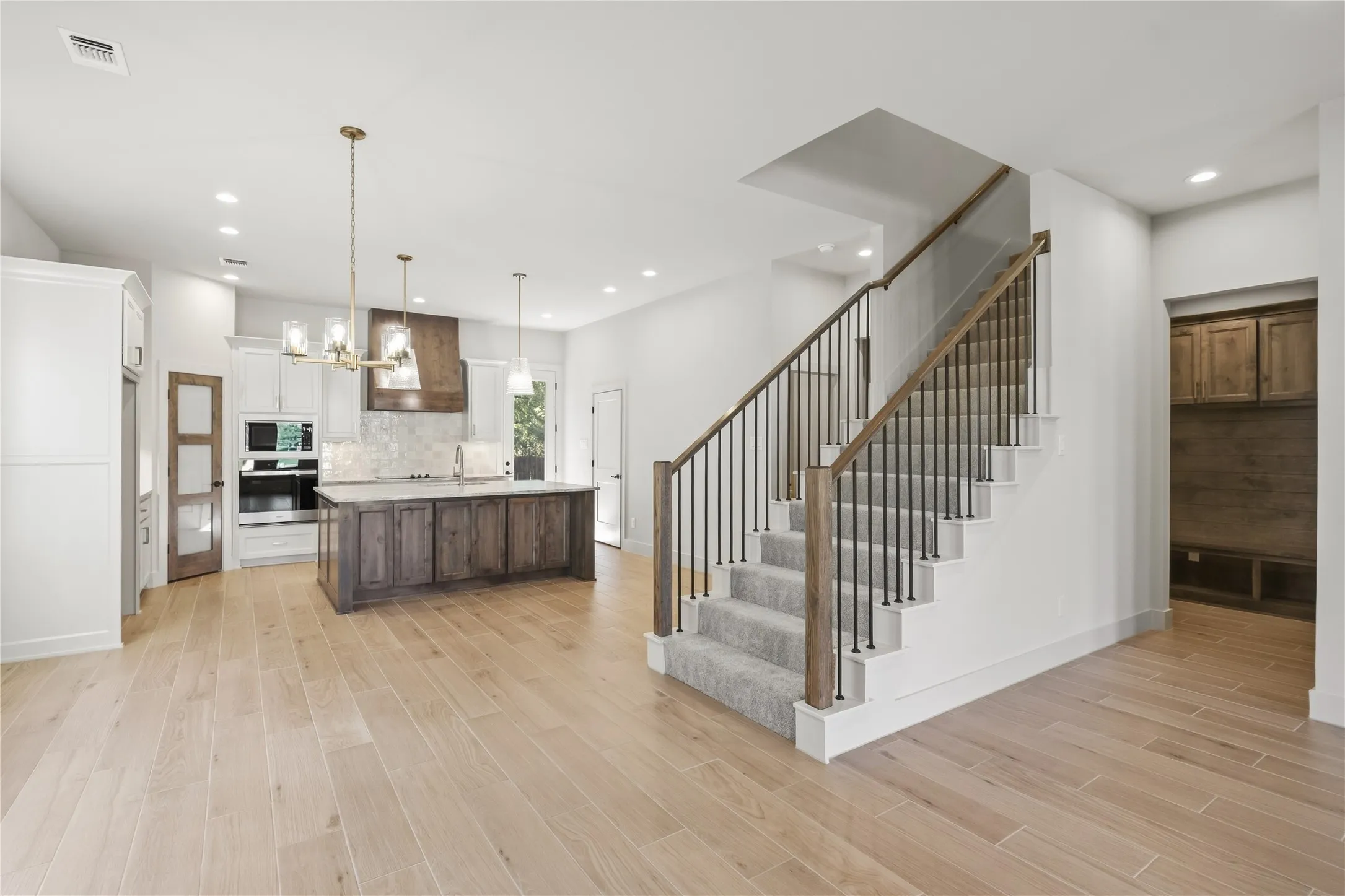 Open-concept interior featuring light wood-finish flooring, a kitchen island with stone countertop, built-in cabinetry, modern staircase with metal balusters, and recessed lighting