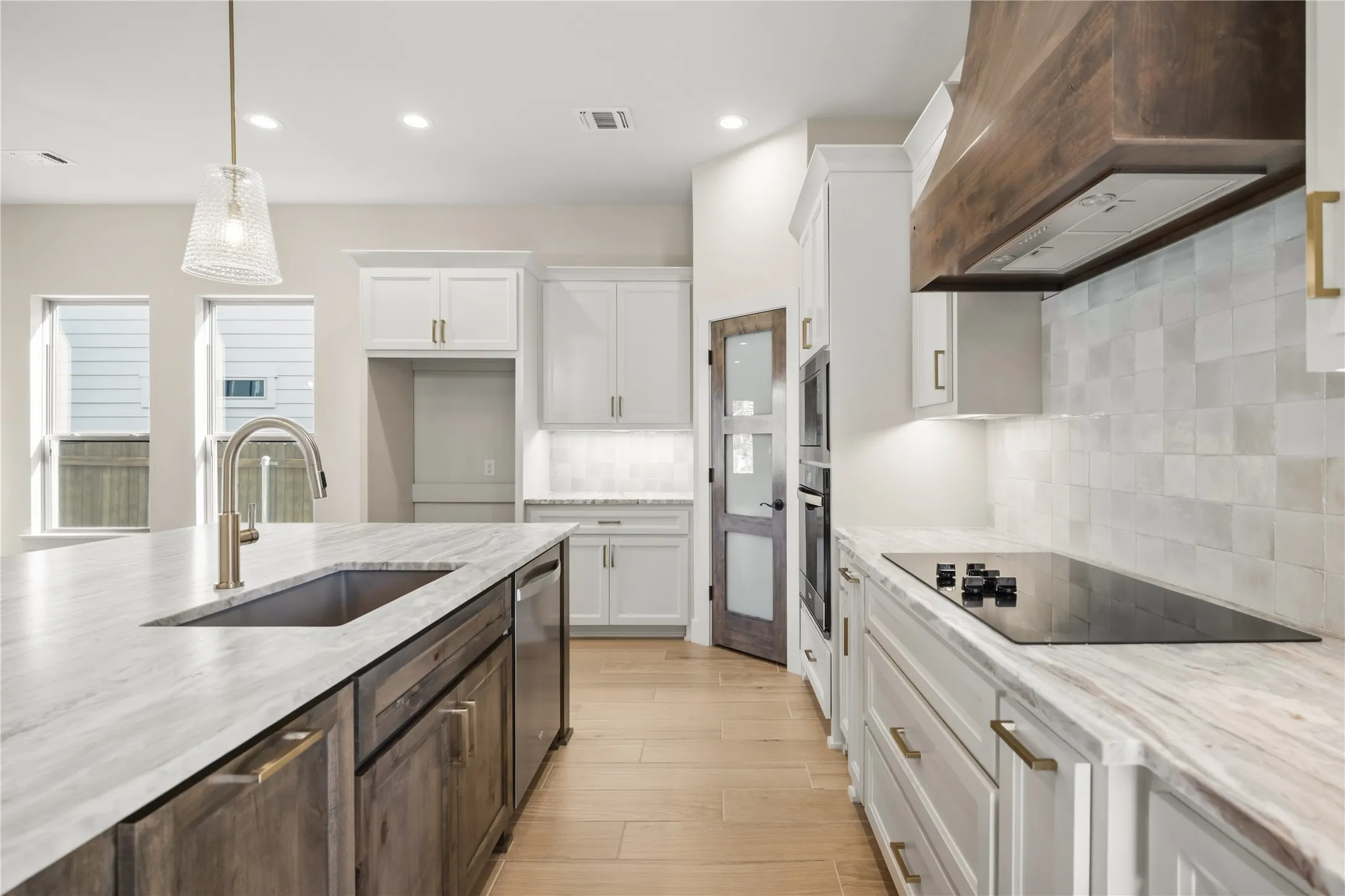 Kitchen featuring an expansive center island with an undermount sink, a wood-finish range hood, white shaker cabinetry, an integrated cooktop, and built-in ovens