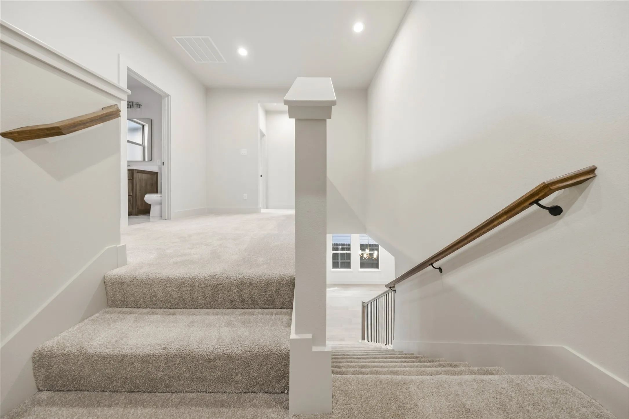Carpeted stairs leading to an upstairs landing with recessed lighting, wood handrails, and a half wall with a white newel post