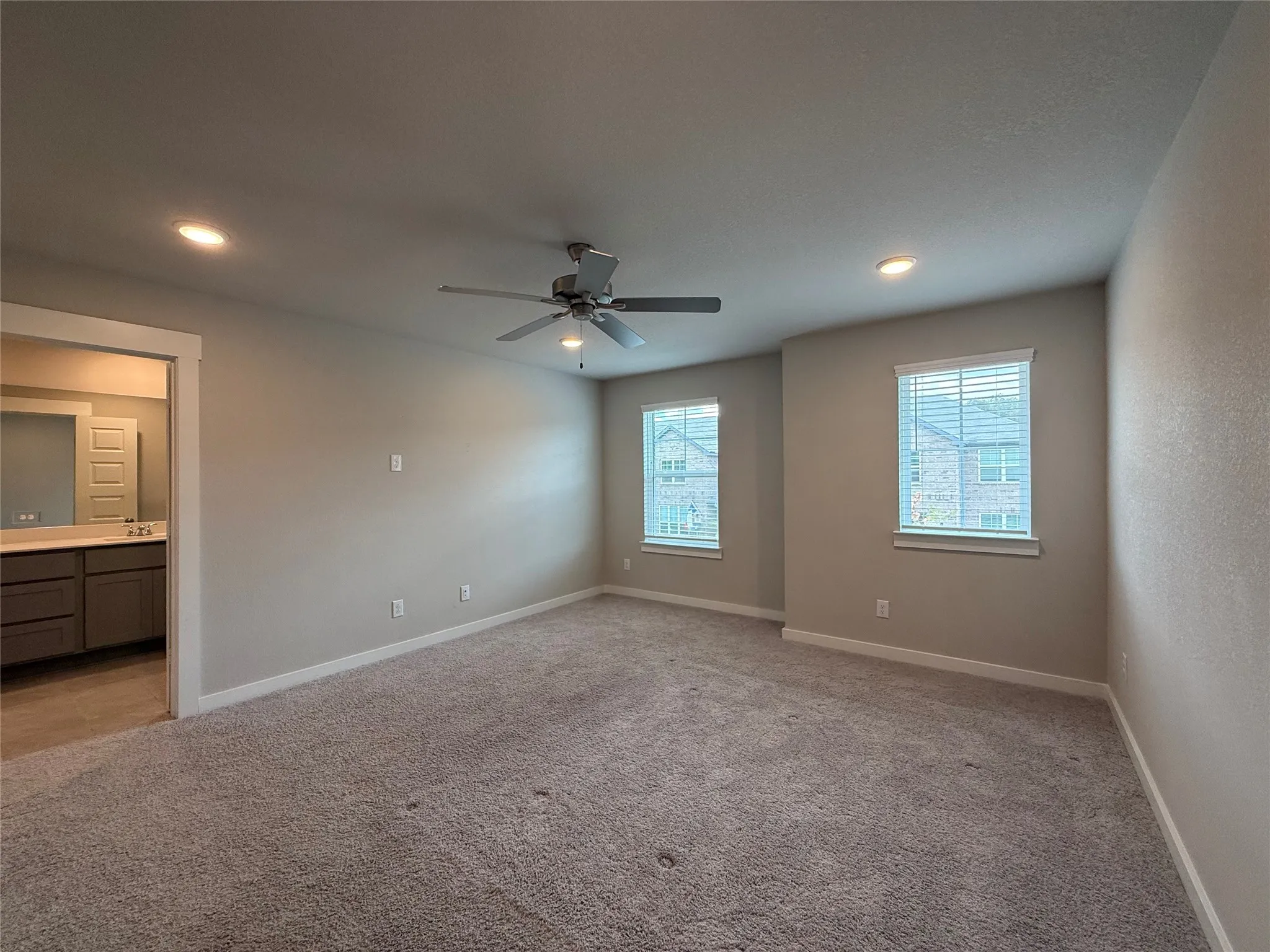 Recessed lighting, ceiling fan, carpeted flooring, two windows with blinds, and an open doorway to a bathroom